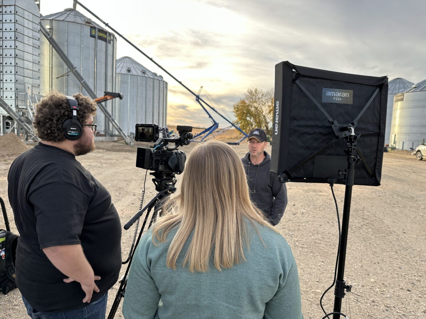 A film crew sets up equipment for a shoot outside in front of several large grain silos. The crew is setting up lighting and a camera as they prepare to film an interview with a man in a baseball cap.