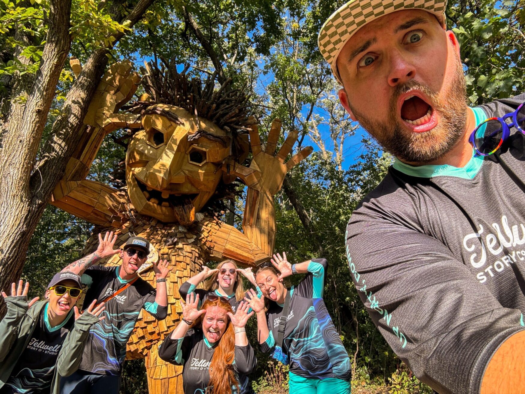 Group selfie in front of a large wooden troll sculpture in a forest. Everyone is making funny faces and hand gestures, matching the troll's playful expression. The group members are wearing Tellwell Story Co. + Studio shirts, and the person taking the selfie is in the foreground with a surprised expression.