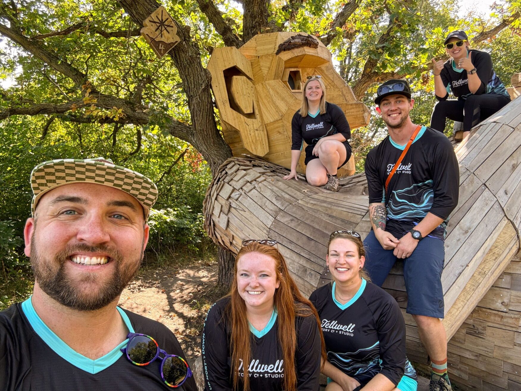 Group of people posing in front of a large wooden troll sculpture, all smiling and wearing Tellwell Story Co. + Studio shirts. One person in the foreground is taking a selfie, and the group is gathered around the troll’s arm, with trees in the background.