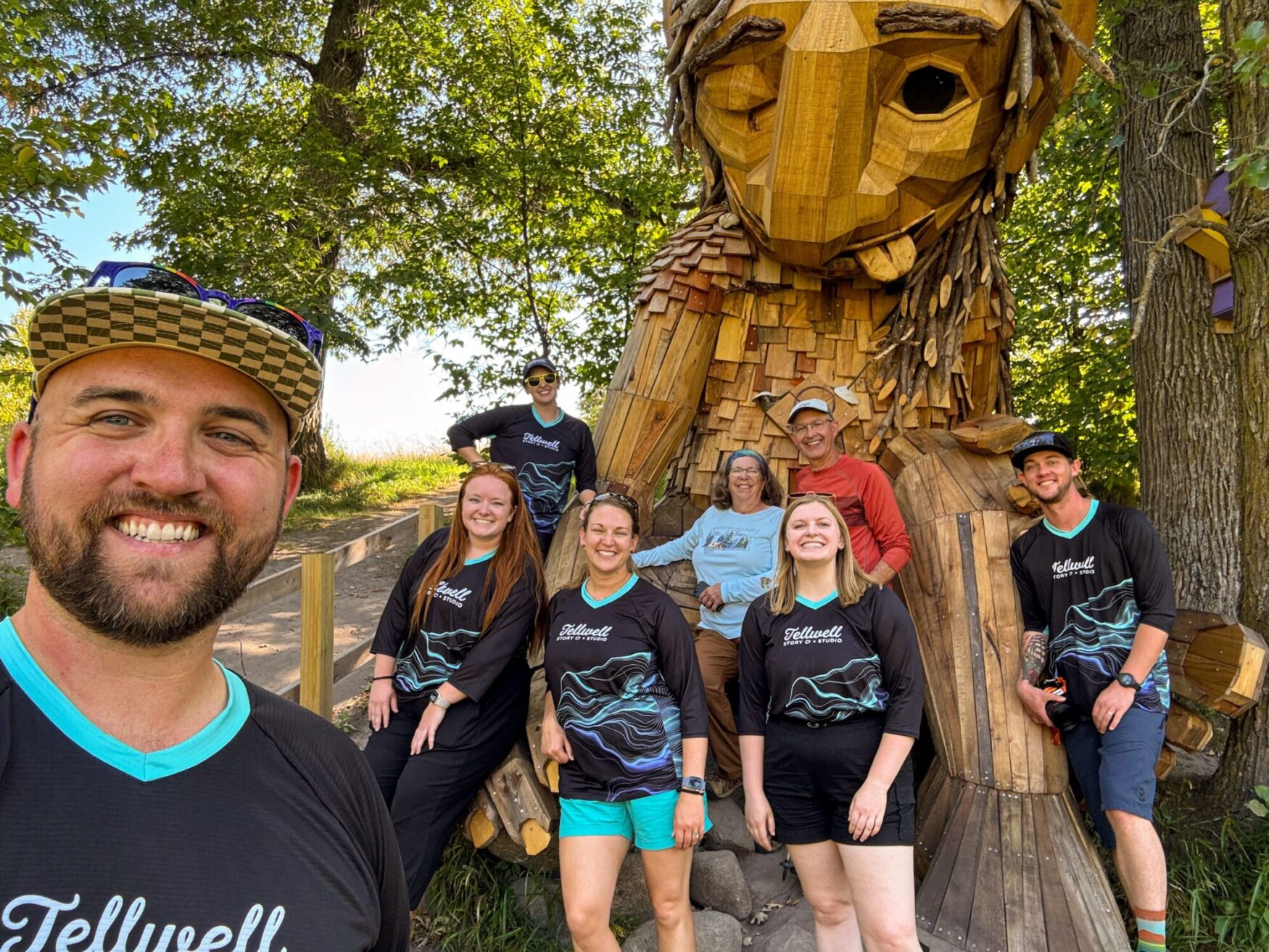 Group of people posing in front of a large wooden troll sculpture, with everyone smiling and wearing Tellwell Story Co. + Studio shirts. The setting is a forested area with trees, and the group is enjoying a sunny day outdoors.