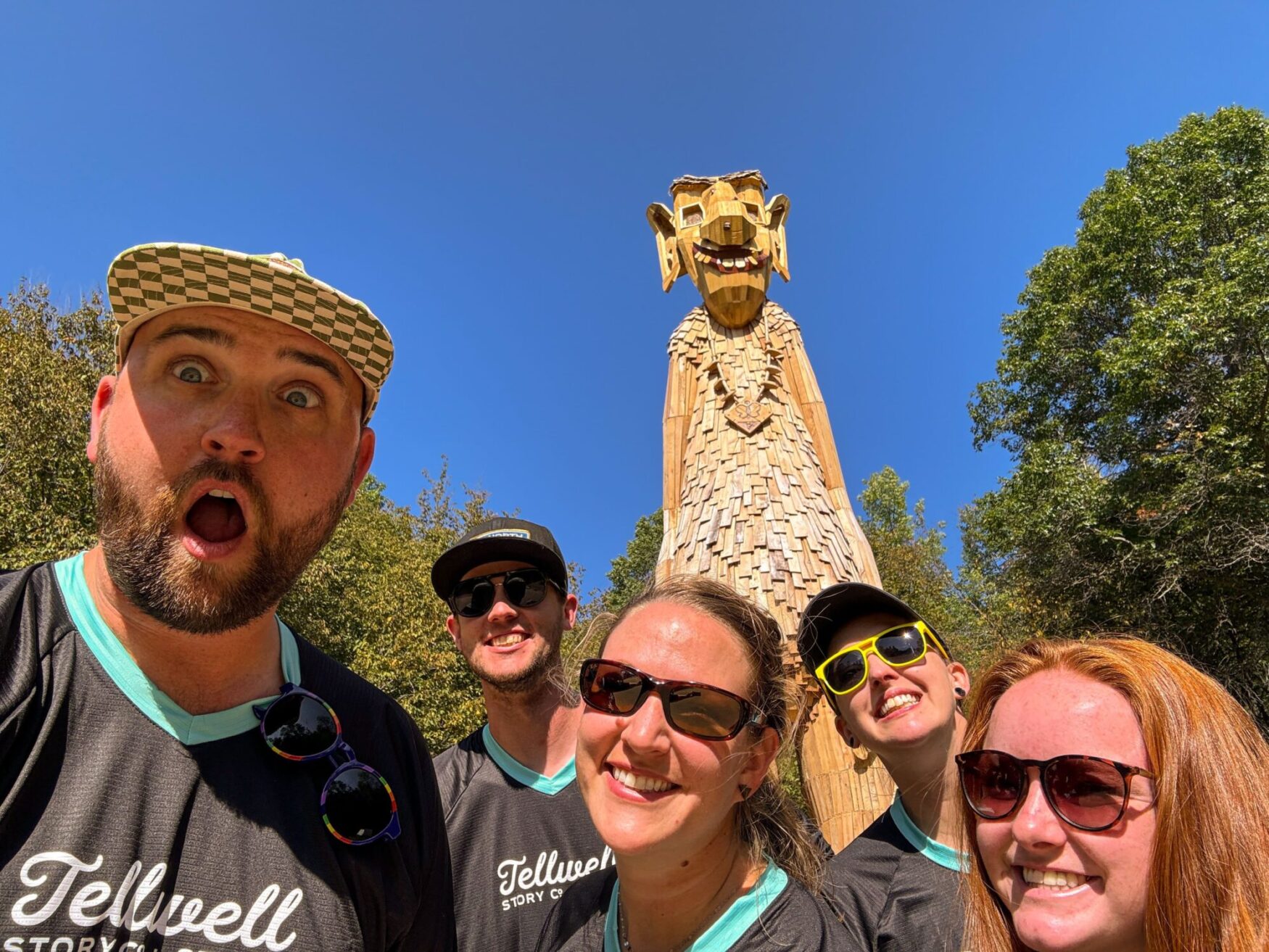 Group selfie with five people wearing Tellwell Story Co. + Studio shirts, posing with excited expressions in front of a tall wooden troll sculpture. The troll and the group are set against a clear blue sky and surrounded by trees.