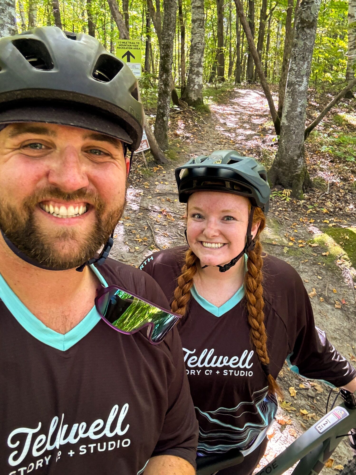 Two people wearing Tellwell Story Co. + Studio shirts and helmets, smiling for a selfie on a wooded trail. They appear ready for a biking adventure, with trees and a trail sign visible in the background.