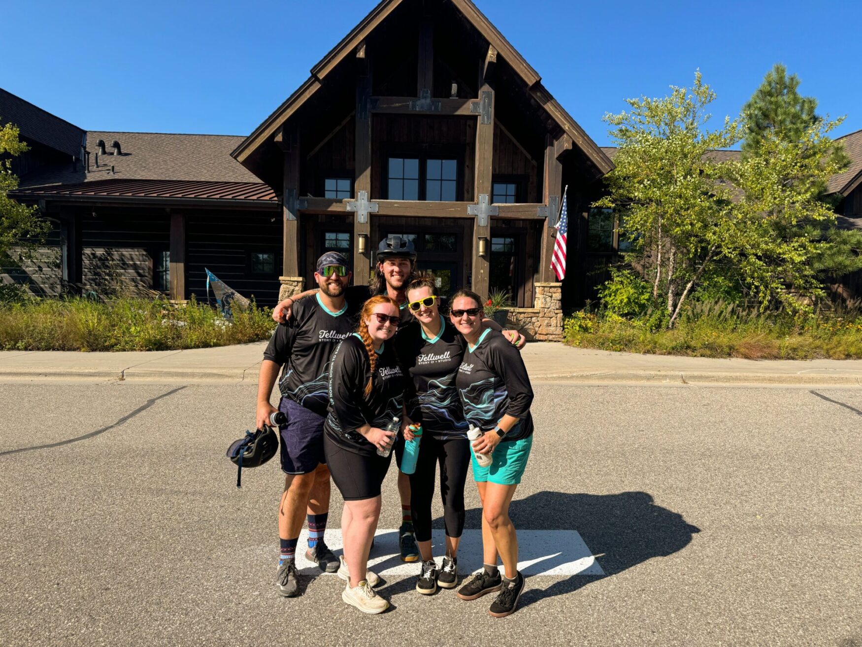 Group of five people wearing Tellwell Story Co. + Studio shirts posing together in front of a rustic building with a wooden facade and an American flag. They are smiling and enjoying a sunny day.