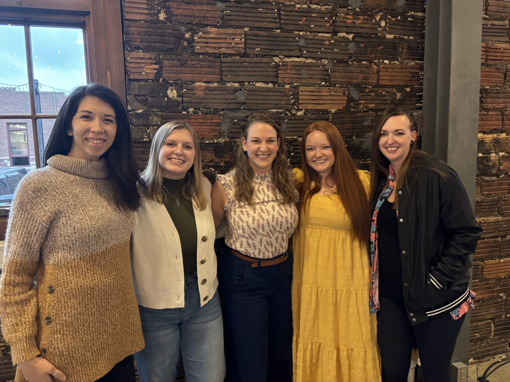 Five women smile and pose together in front of a rustic brick wall. They are dressed warmly, with one woman wearing a long yellow dress. The group looks cheerful as they stand closely side by side.