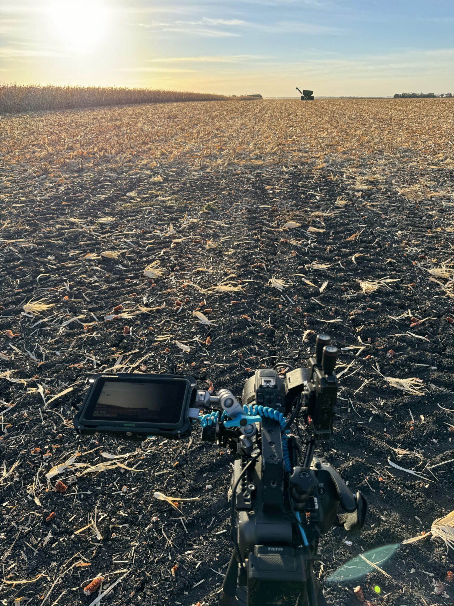 In this photo, a professional camera is resting on the ground on what appears to be a farm field. A tractor is harvesting corn in the background. It is sunset, which showcases the yellow fields and blue sky.