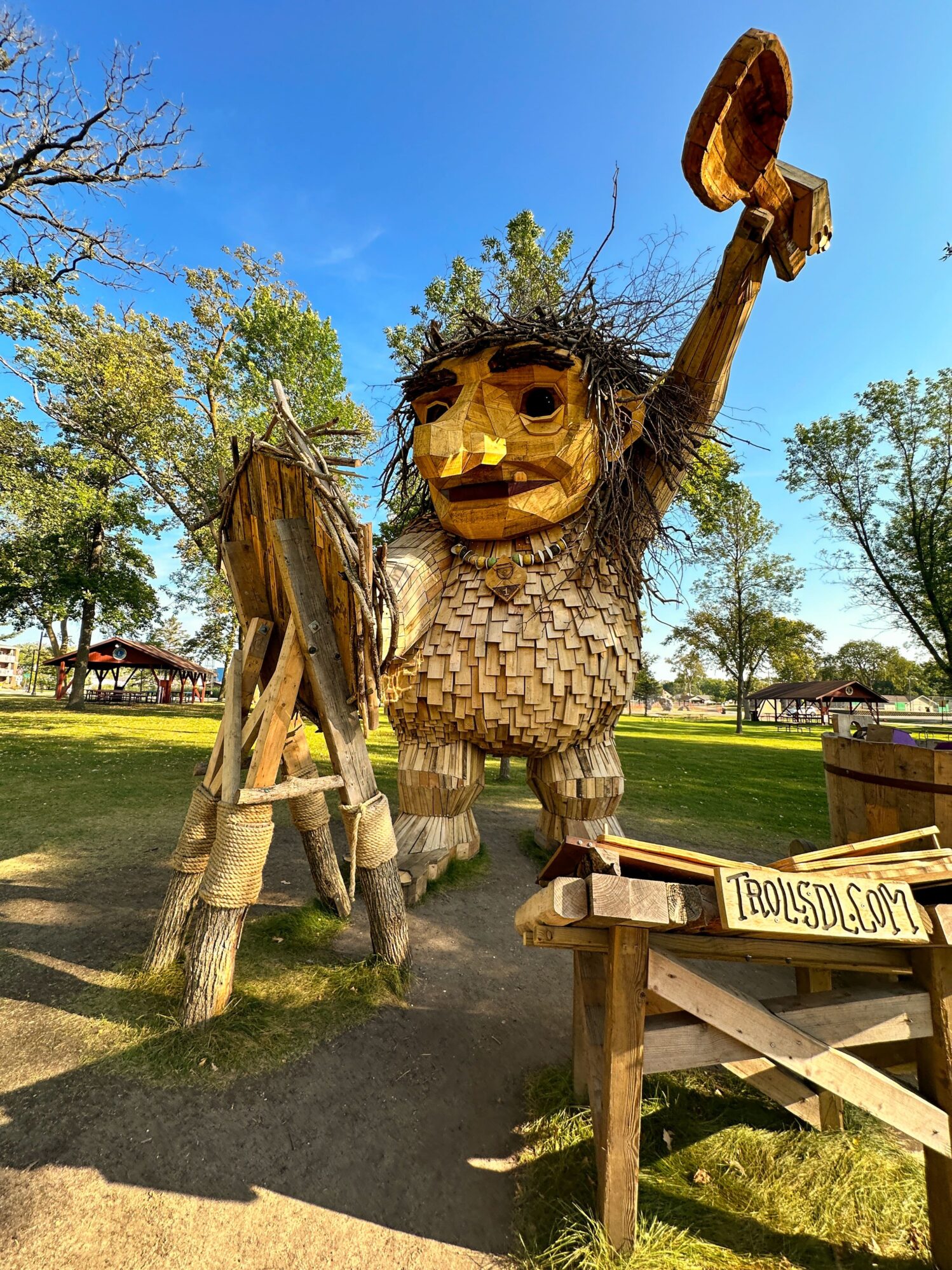 Large wooden troll sculpture standing in a park, holding a tree stump in one hand and a birdhouse in the other. A wooden sign in the foreground reads 'TrollsDL.com.' The setting includes trees and open green spaces under a clear blue sky.