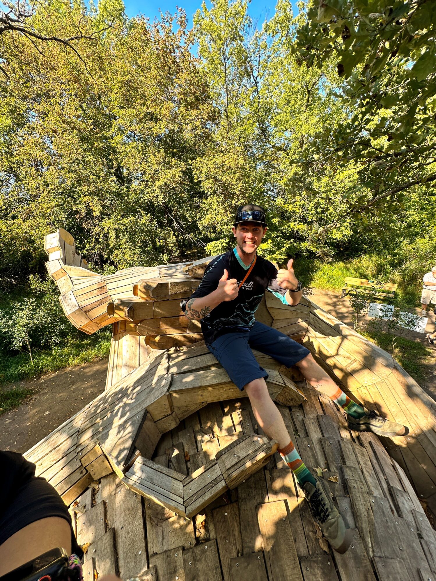 Person sitting on a large wooden troll sculpture, giving a thumbs-up and smiling. They are wearing a Tellwell Story Co. + Studio shirt, surrounded by trees and enjoying a sunny day outdoors.