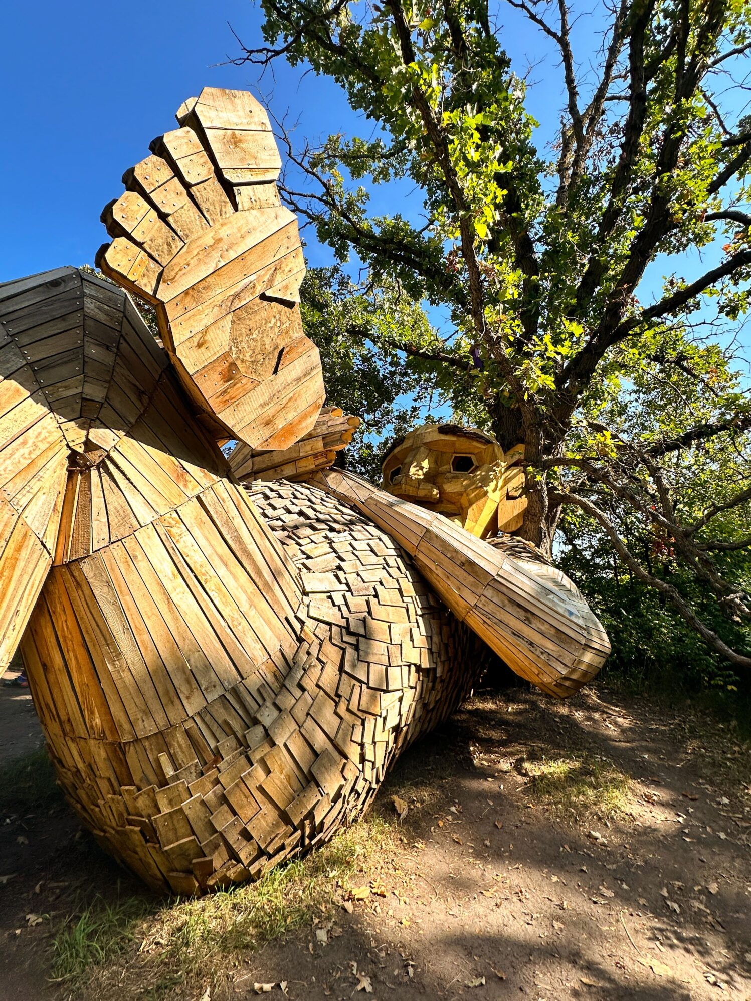 Close-up of a large wooden troll sculpture lying on the ground with its arm extended, surrounded by trees under a clear blue sky. The troll is crafted from numerous wooden planks, giving it a textured, layered appearance.