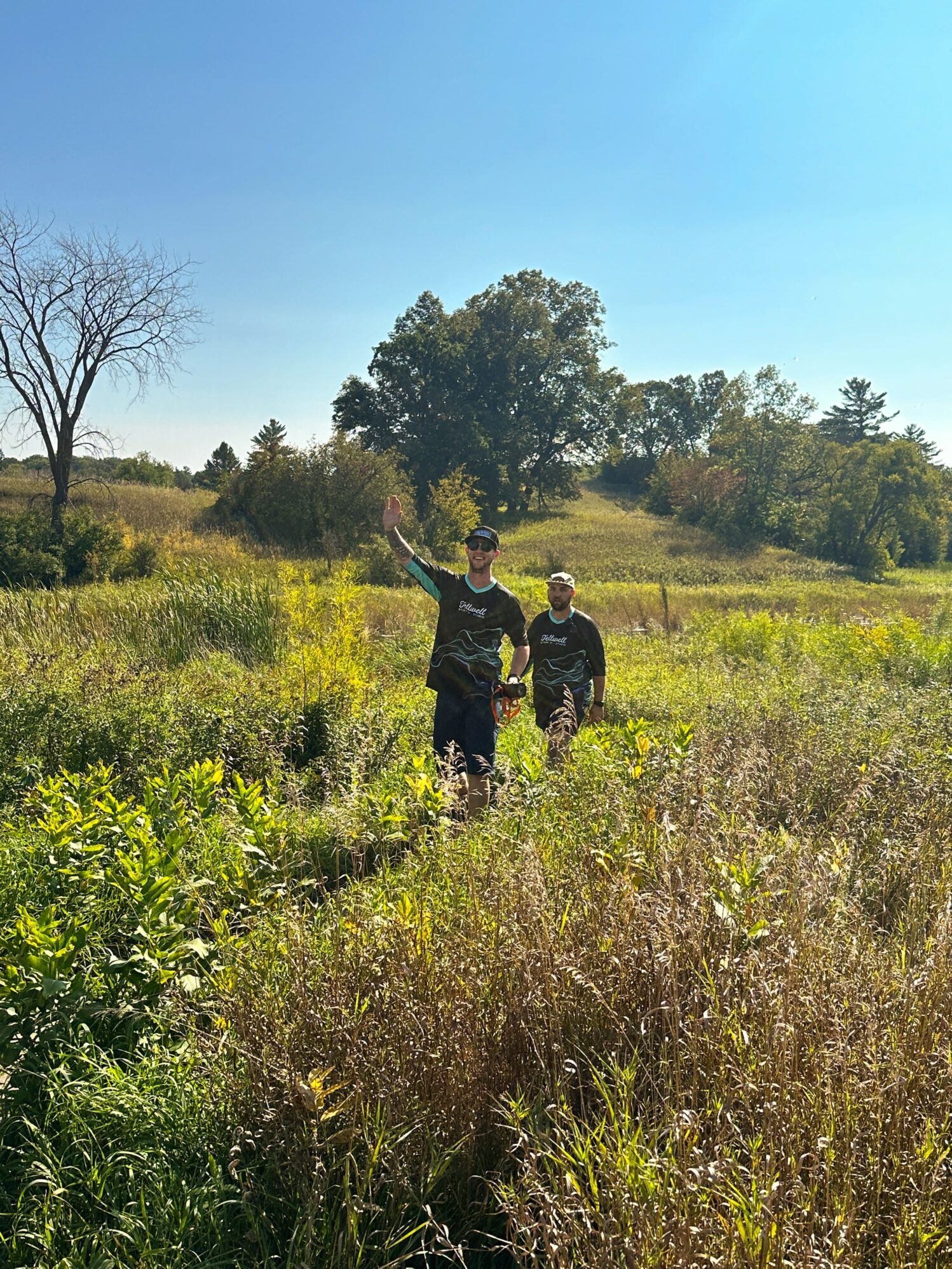 Two people walking through a field of tall grass and wild plants on a sunny day. One person is waving, and both are wearing Tellwell Story Co. + Studio shirts. The background includes rolling hills and scattered trees under a clear blue sky.