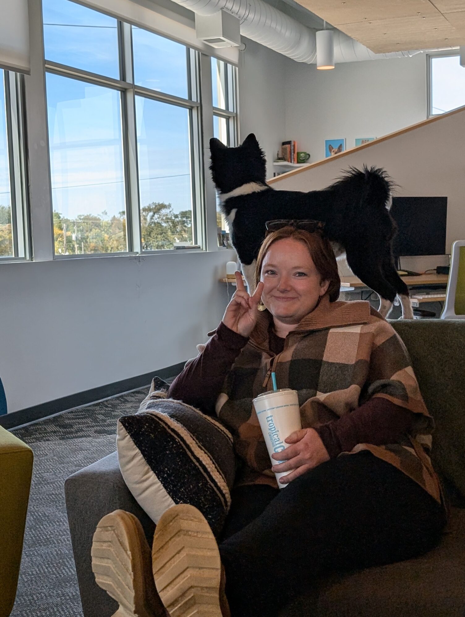 A woman sitting on a couch, smiling and holding a drink with a 'Tropical Smoothie' cup, flashing a peace sign. A black and white dog stands on the couch behind her, looking out large windows with a view of trees.