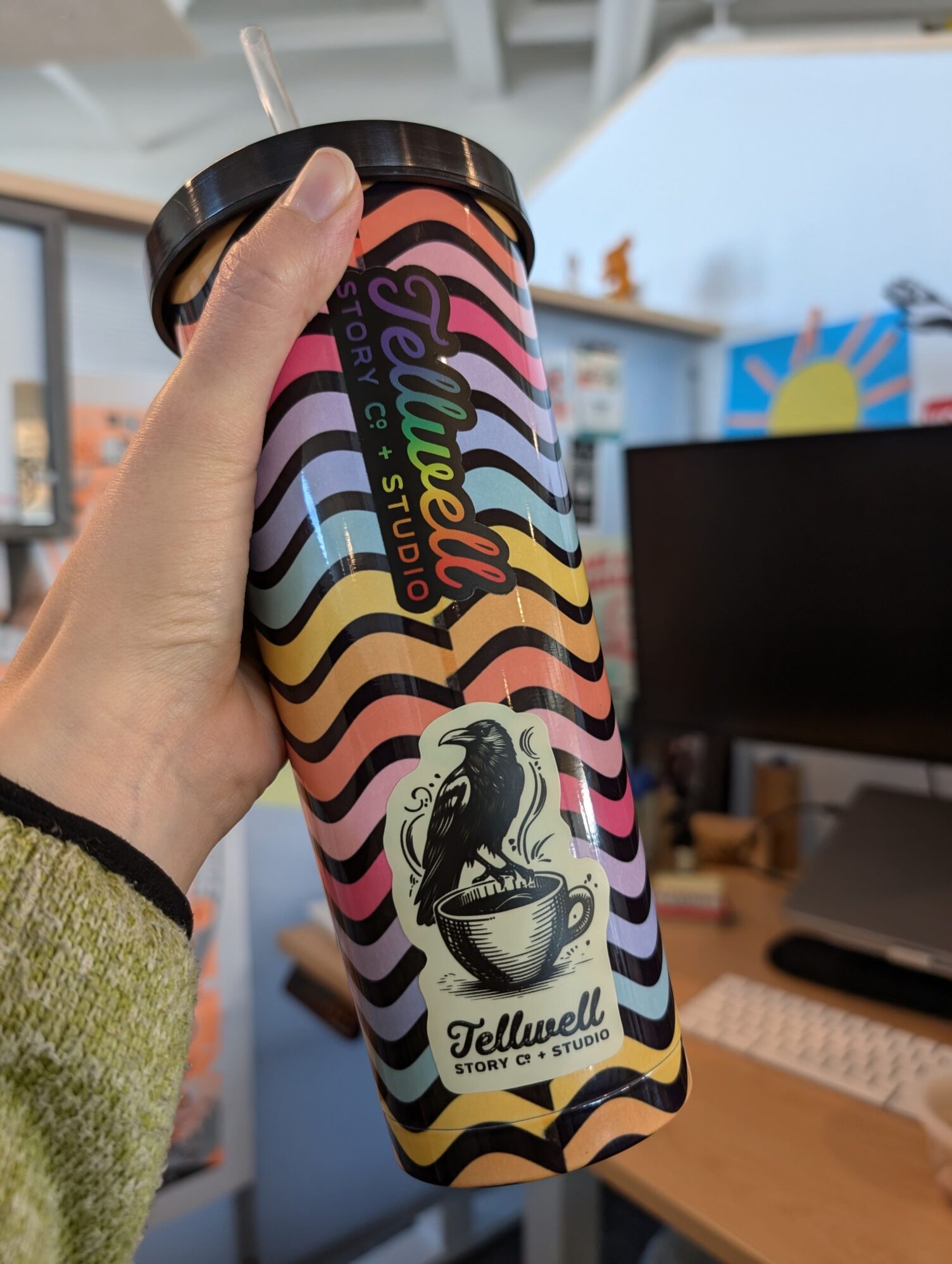 Hand holding a colorful tumbler with wavy rainbow stripes and the Tellwell Story Co. + Studio logo, featuring a black crow sitting on the edge of a coffee cup. Office space with a computer monitor and desk in the background.