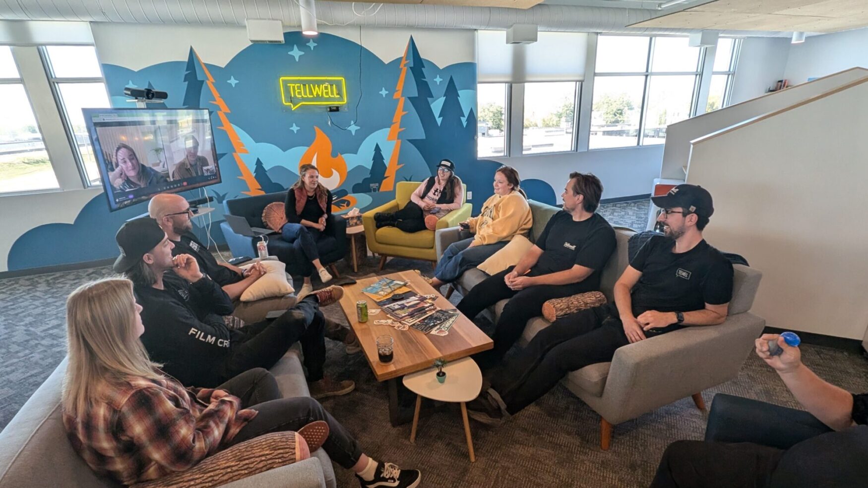 A group of people sitting on couches positioned in a circle, all facing each other, including a TV screen on wheels with someone joining virtually, all with a coffee table at the center. The atmosphere is pleasant, and everyone is engaged in conversation.