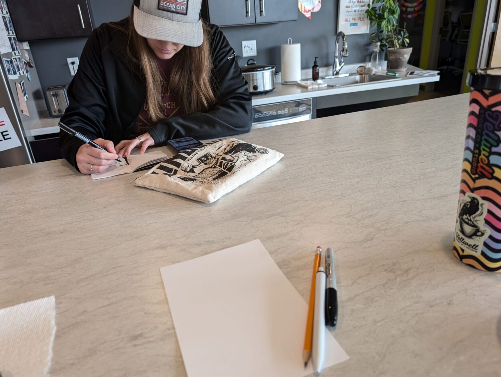 A woman sitting at a table, head down, with a hat on, drawing in a sketchbook with a bag of markers in front of her. In the foreground is a sheet of blank paper with pens and a pencil sitting atop.