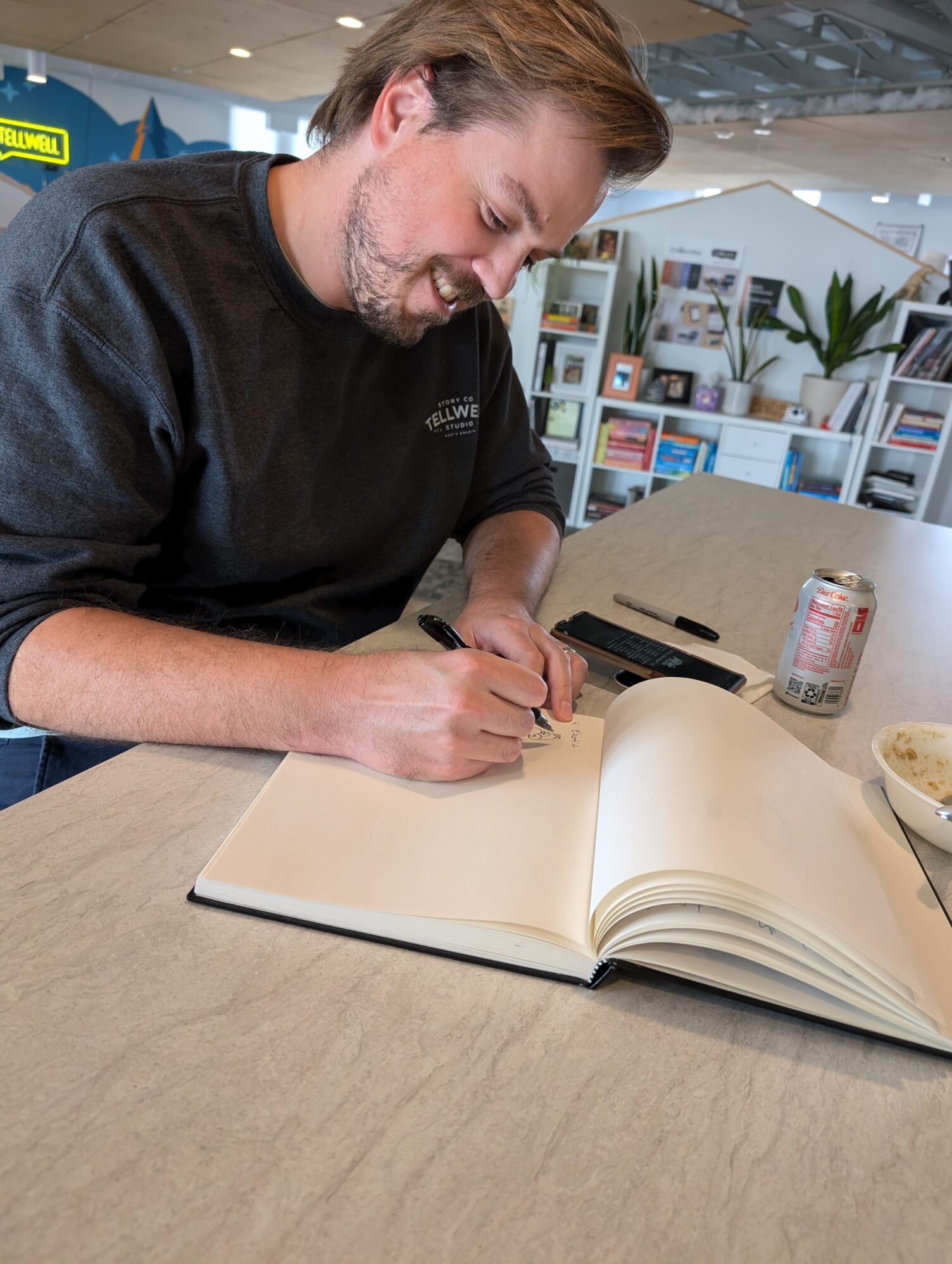A man smiling and sitting at a table while drawing and writing something in an unlined sketchbook with a pen. Next to him are a cell phone, another pen, a drink, and a bowl. A bookcase in the background.