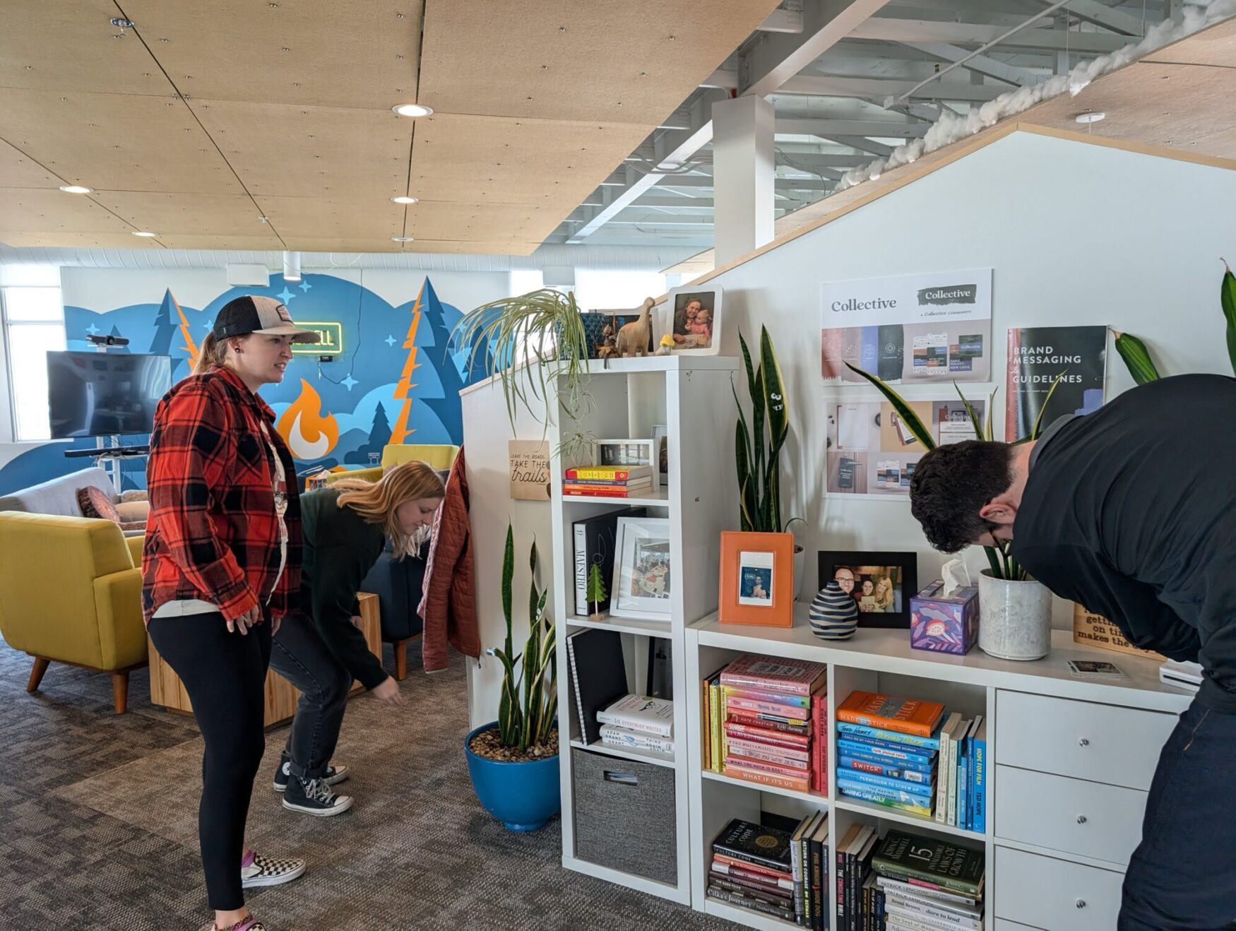 Three coworkers in casual attire stand around a bookshelf in a brightly lit office. One person is leaning down, another is bending over the shelf, and a third person is smiling, observing the scene. The room is filled with books, plants, and office decorations.