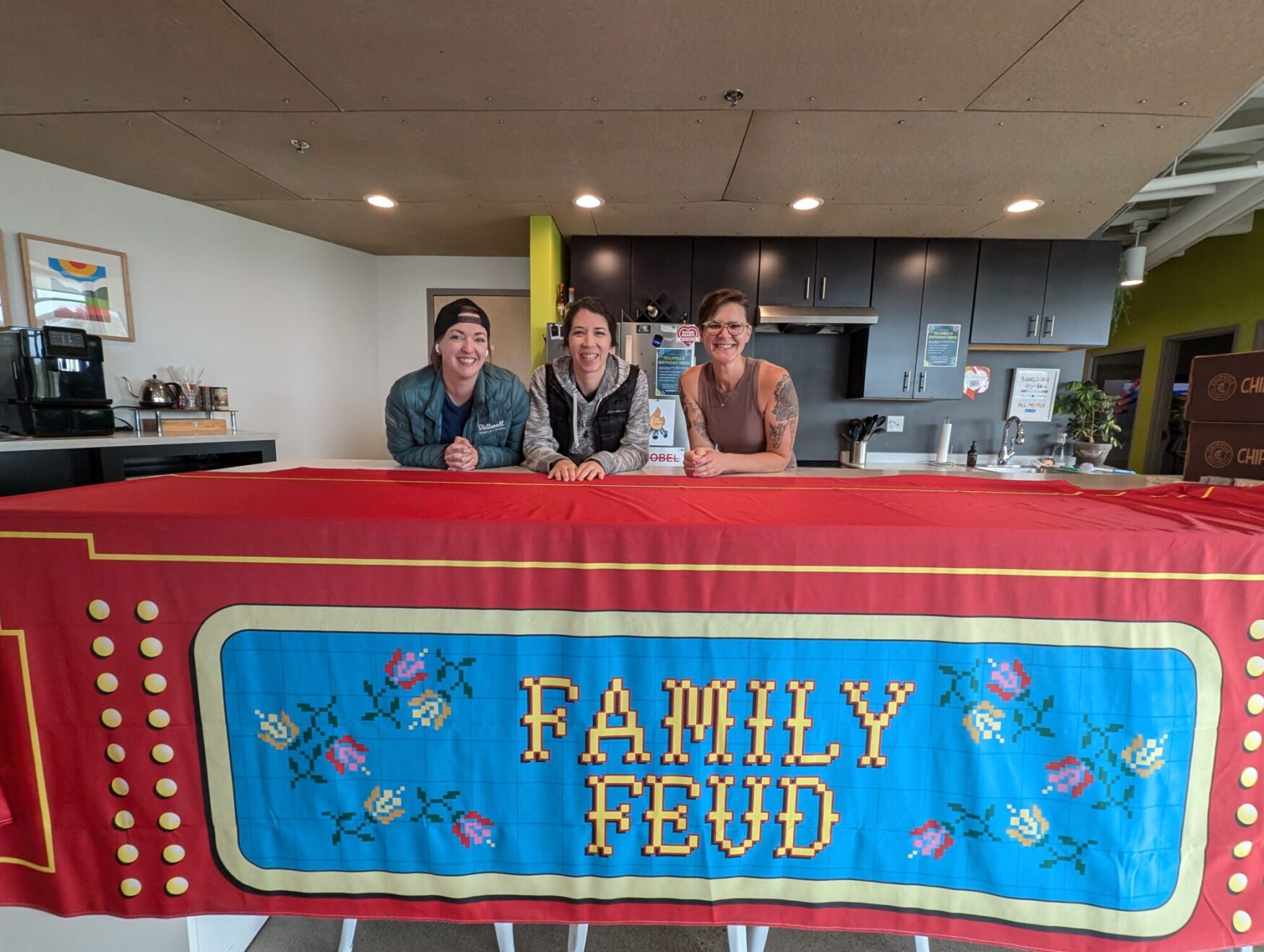 Three people standing behind a counter covered with a large red banner that resembles the design of the game show Family Feud. The banner has a retro cross-stitch floral pattern and bold yellow lettering spelling 'Family Feud.' The individuals behind the counter are smiling, with one holding a cup, in a kitchen area with cabinets, a coffee machine, and various supplies in the background.