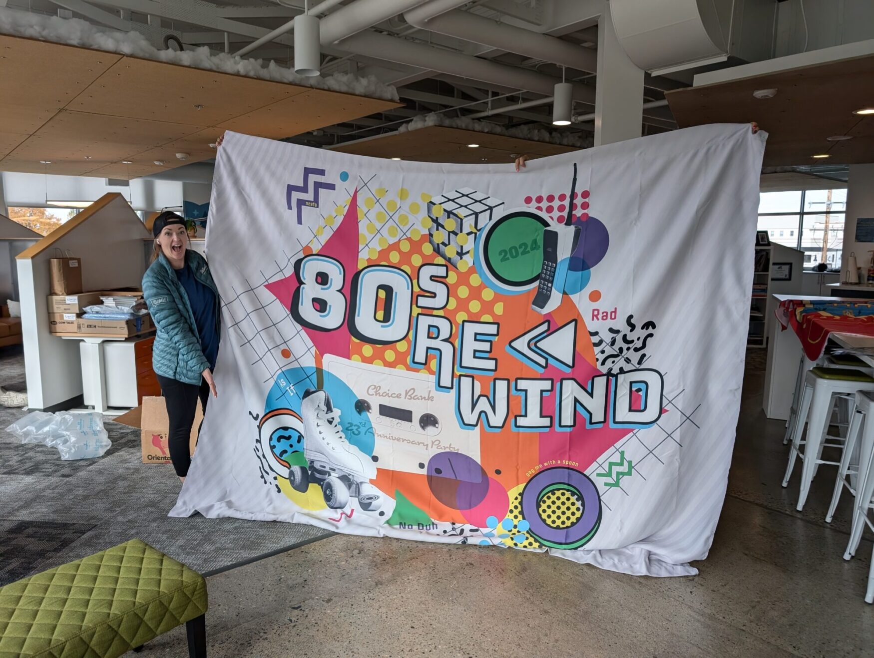 A person excitedly holds up a large banner with a colorful 80s-themed design that reads '80s Rewind.' The banner features retro graphics, including a Rubik's cube, a cassette tape, a roller skate, and an old-school cell phone, along with vibrant colors and geometric patterns. The person is wearing a teal jacket and a backwards cap, posing in a modern office space.