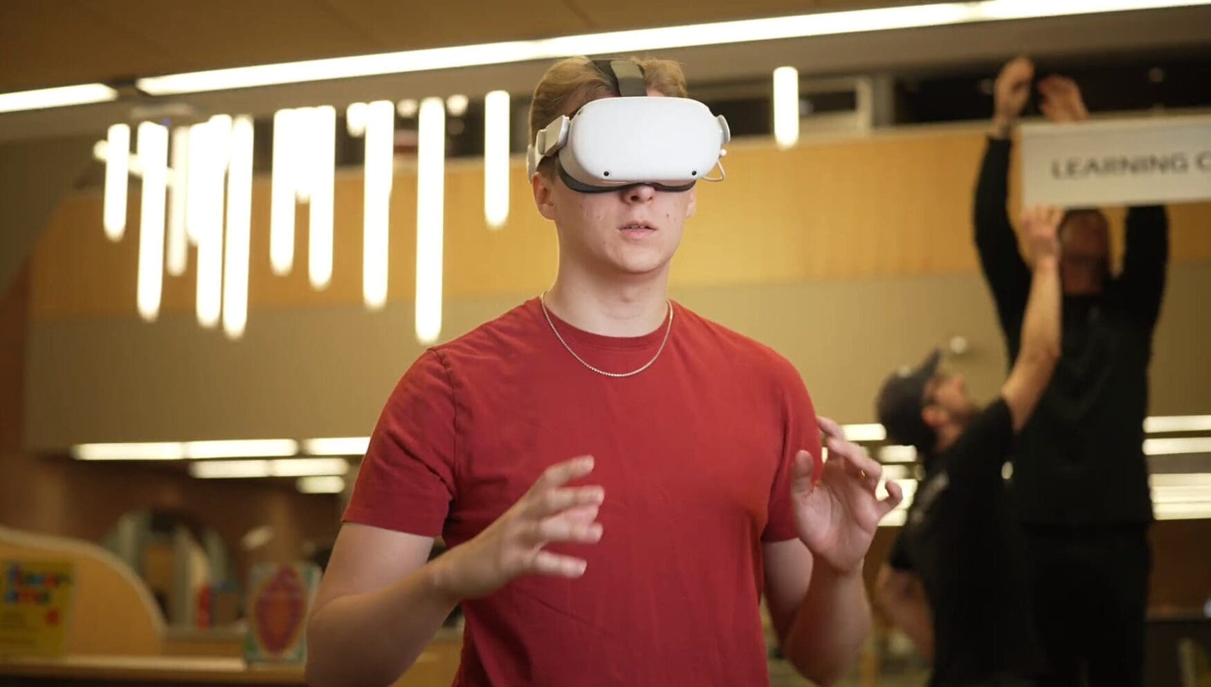 A young man in red wearing a virtual reality headset stands in a brightly lit room, appearing focused with his hands raised slightly.
