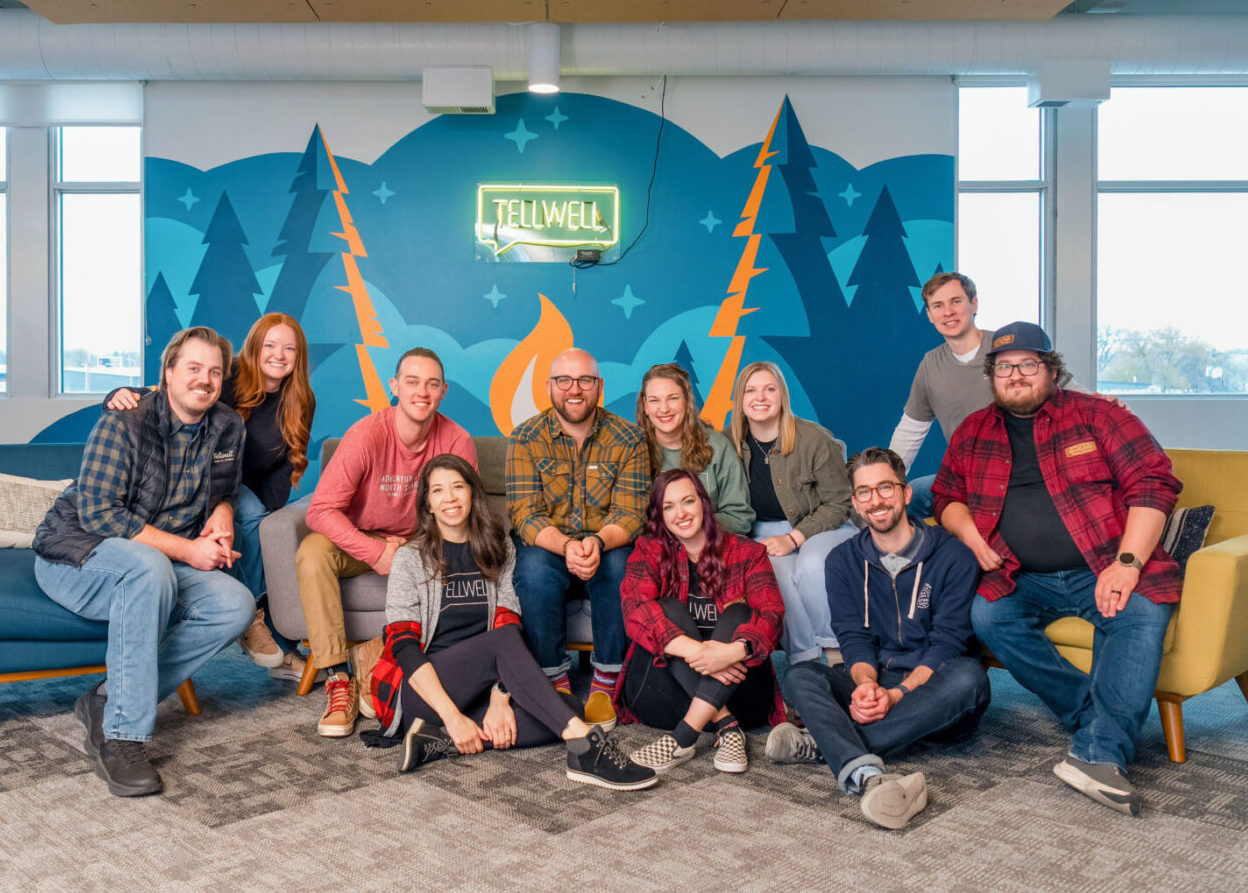 Team photo taken in a modern office space with a colorful mural of trees, mountains, and a campfire with the Tellwell logo in neon. The group is seated and kneeling on sofas and the floor, smiling and casually dressed, giving a warm, friendly vibe.