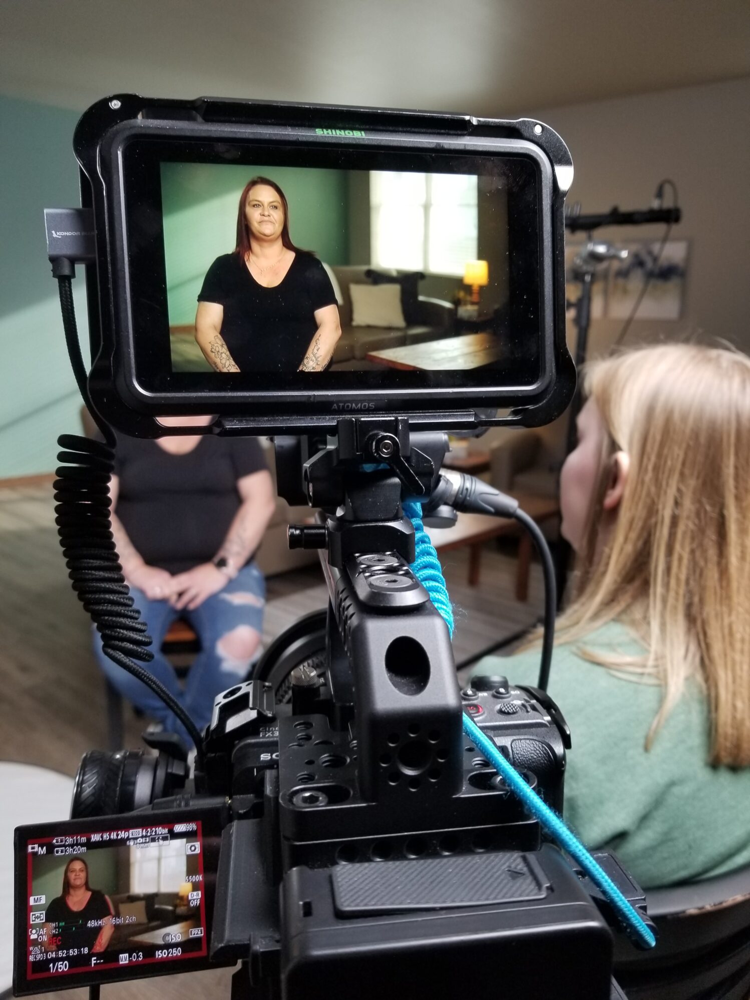 In the picture we see a camera pointed at a woman being interviewed. To the right of the camera someone is conducting the interview.