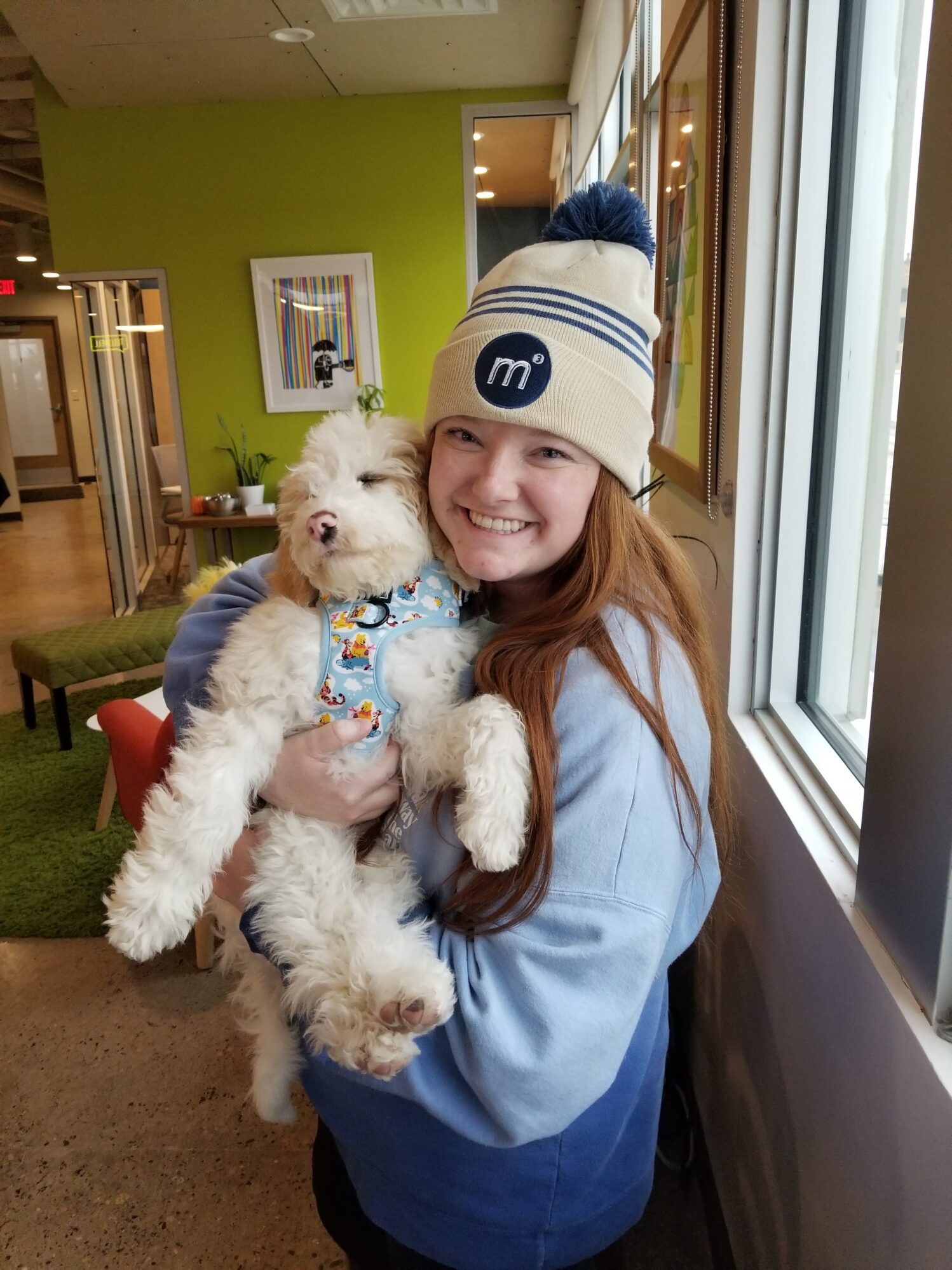 A woman is smiling and holding a cute dog. The dog has white hair and a pink nose. The woman is wearing a beige and blue stocking cap and a light blue sweat shirt.