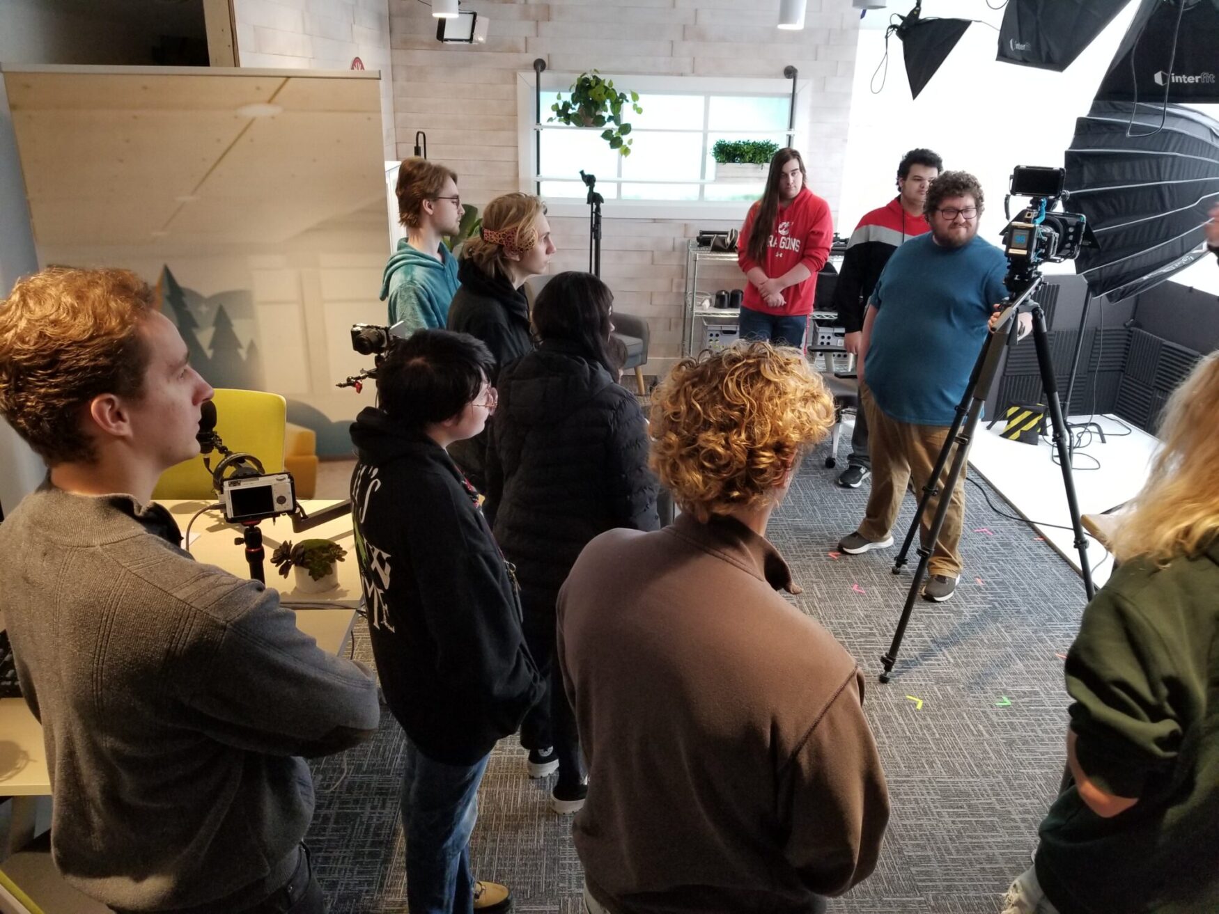 A group of people standing in a studio setting, focused on a film production setup. Cameras, tripods, and lighting equipment are visible, with individuals engaged in discussion or observation.