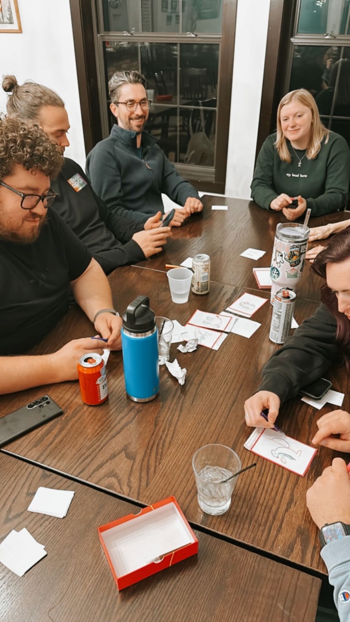 Five people around a table playing card games. There are cups and cans on the table, which appears to create a party atmosphere. They are smiling and having a fun time playing the game.