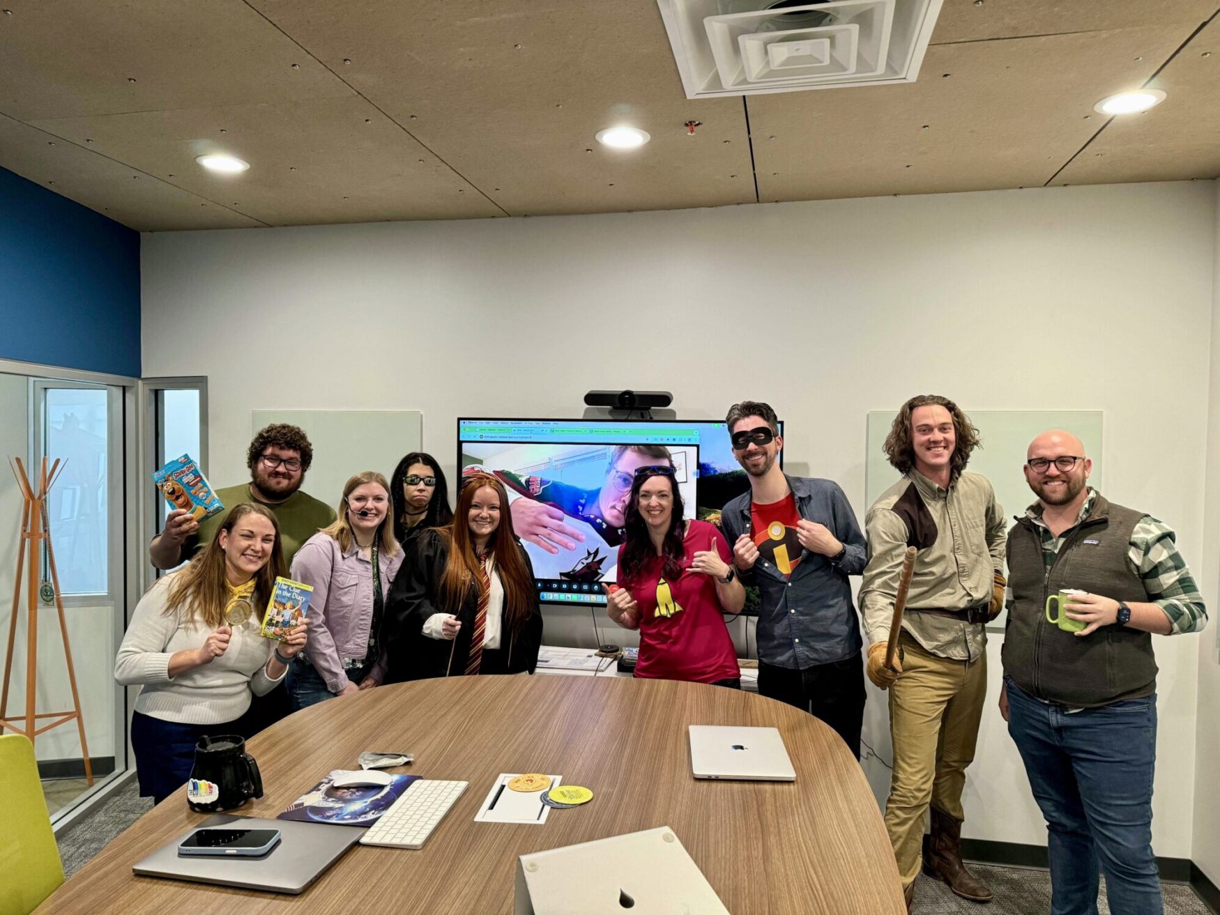 Team photo of people dressed in various costumes, standing in a conference room around a wooden table with laptops and mugs. Some are holding books, a coffee pot, and wearing accessories like superhero masks and wands, giving a fun, lighthearted vibe. A screen behind them displays a video call with another person joining in virtually.