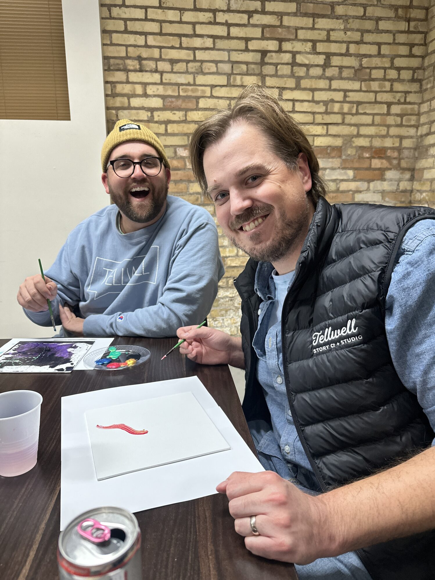 Two men smiling at the camera while painting. They are wearing Tellwell apparel and appear cheerful.