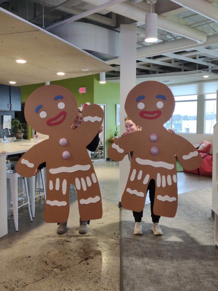 Two oversized gingerbread men cutouts held playfully by team members peeking out from behind them in a brightly lit, modern office space.