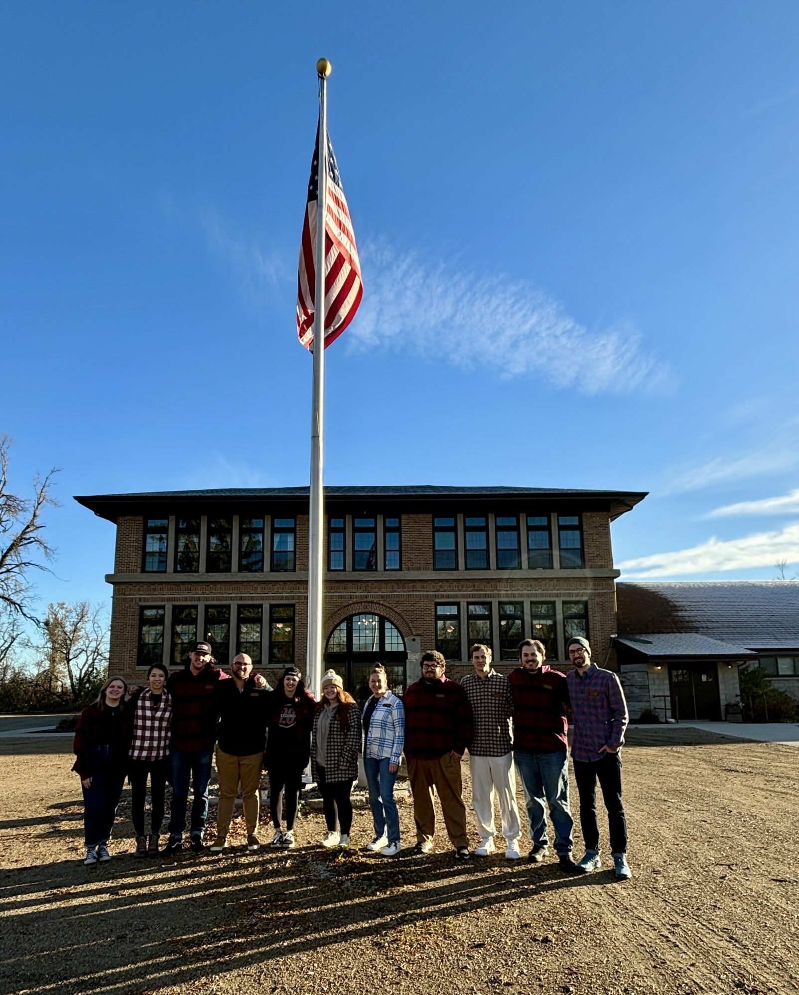A group of people stands in front of a flagpole with the American flag, outside a two-story brick building on a clear day.