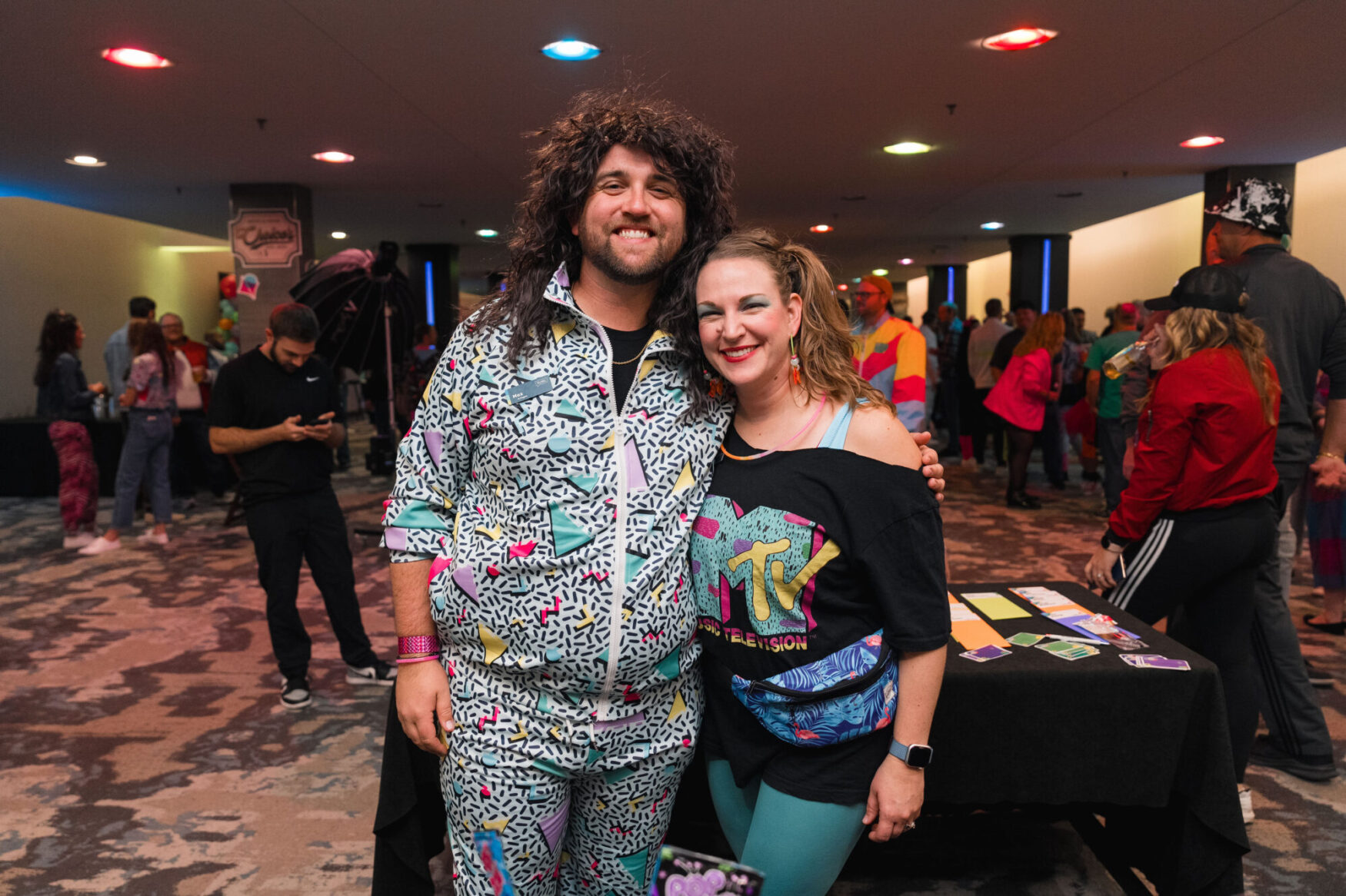 A smiling man and woman in retro 80s outfits stand together in a lively indoor event space.
