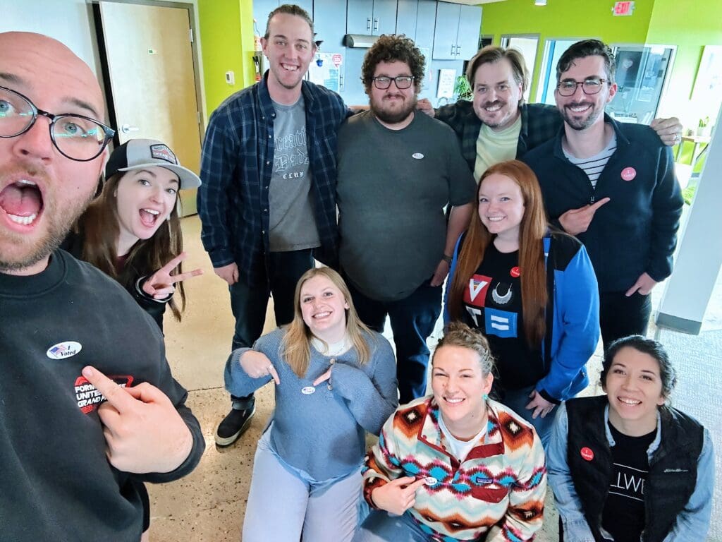 This group photo shows ten smiling people in a bright room with lime-green walls, creating an energetic atmosphere. Most of them are wearing "I Voted" stickers, indicating they recently participated in an election. In the front, a few people point to their stickers, adding to the playful vibe. The group appears cheerful and engaged, with everyone smiling for the camera. The room’s vibrant decor enhances the positive, upbeat energy of the photo.