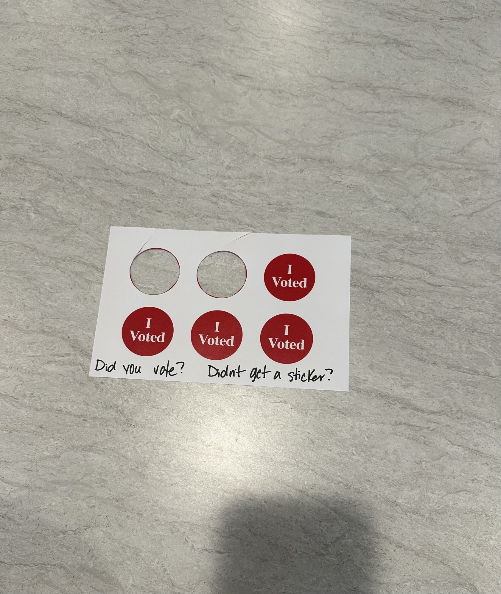 A sheet with red "I Voted" stickers lies on a light gray surface. Two stickers have been removed, leaving empty circles, while four stickers remain. Below the stickers, handwritten text reads, "Did you vote? Didn't get a sticker?"