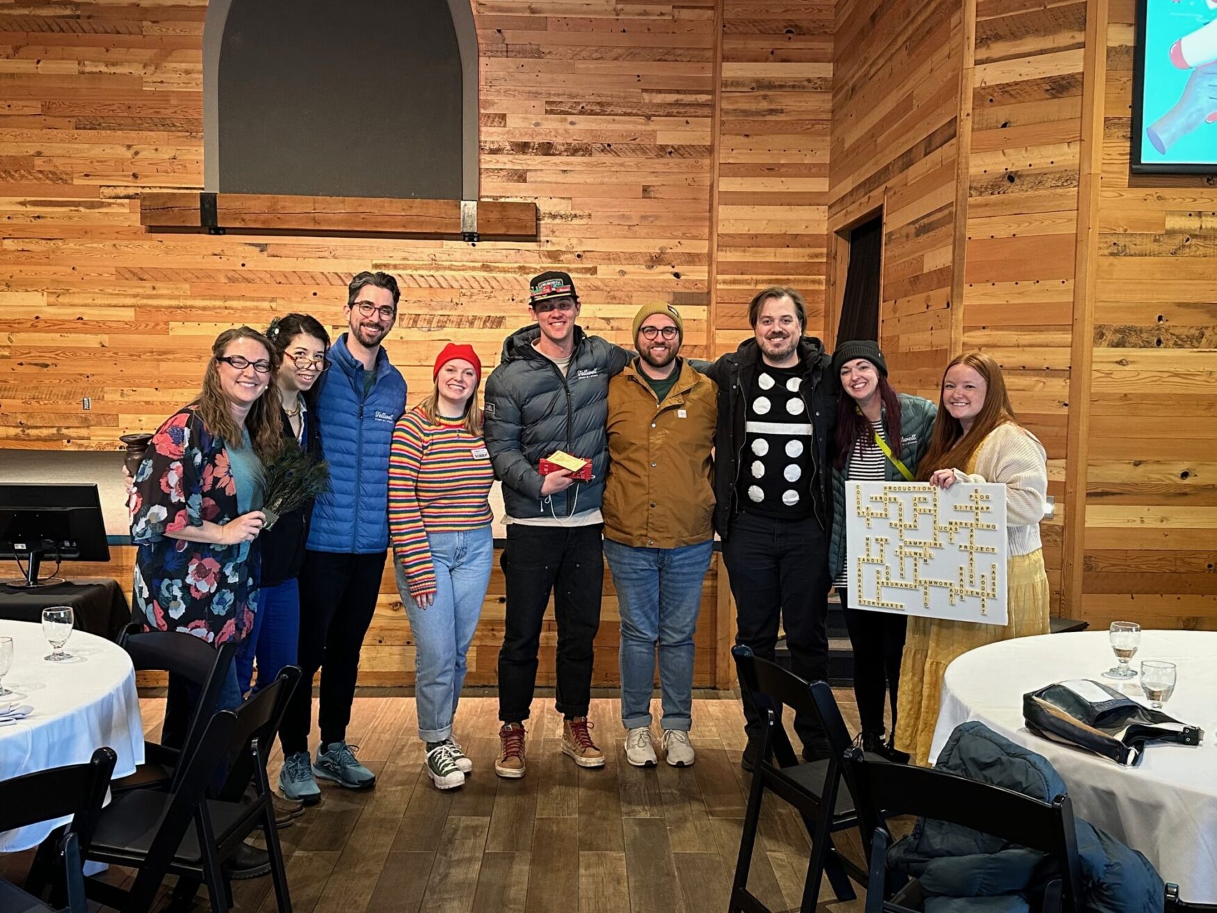 Group photo of Tellwell Story Co. + Studio team members standing together in a warmly lit, wood-paneled room. Several team members are wearing costumes or holding props, including a Scrabble board and a fan of peacock feathers. The group is casually dressed, smiling, and standing in front of a wall with rustic wood paneling.