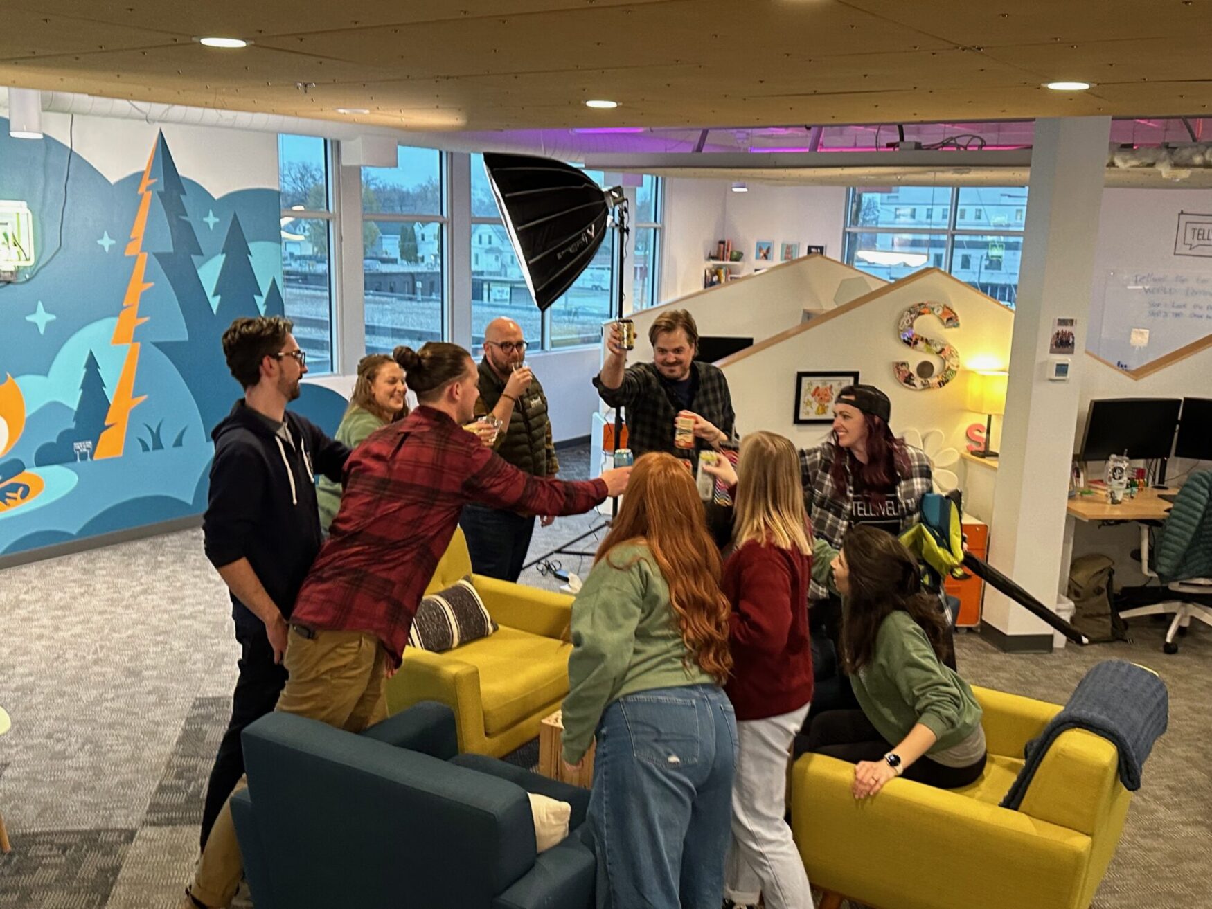 A group of people gathered in an open, colorful office space with large windows. They are standing or sitting around a set of yellow and blue chairs, raising their drinks together in a cheerful toast. The walls are decorated with bright, playful artwork featuring trees and abstract shapes, and there's a large 'S' on the wall adorned with small photos. A photography light is visible in the background, adding to the vibrant and lively atmosphere.