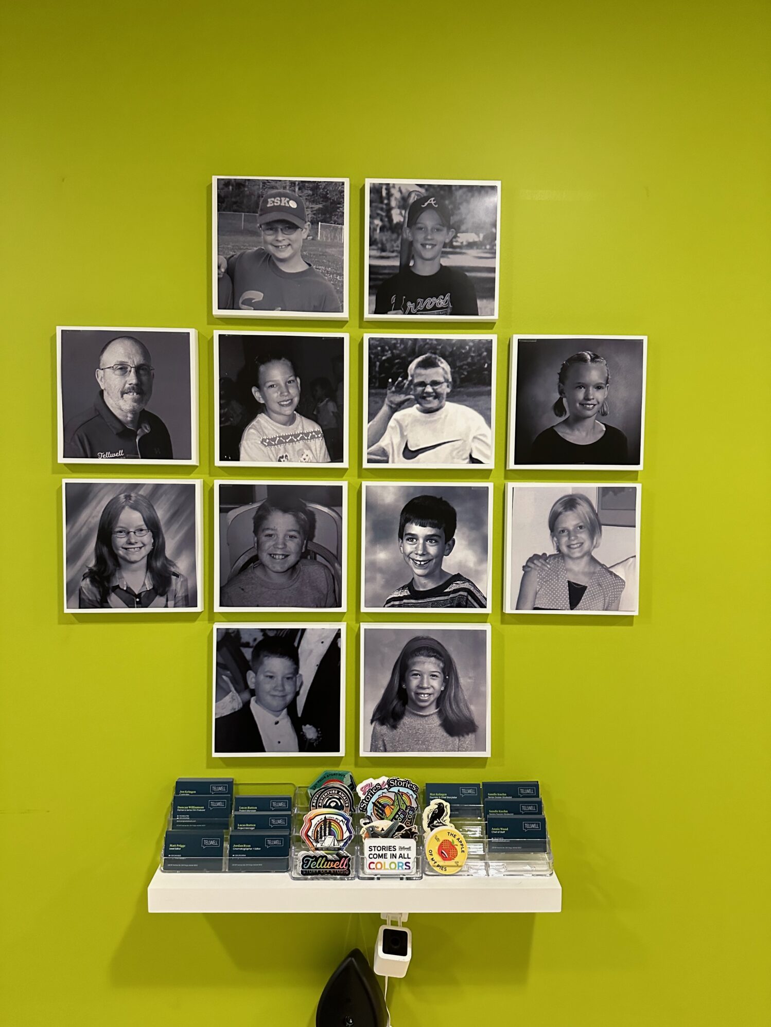 A lime green wall featuring 12 headshots. Each headshot is of a child, roughly at 10 years old. There are 7 men and 5 women pictured. Below the photos is a small shelf that holds stickers and business cards.
