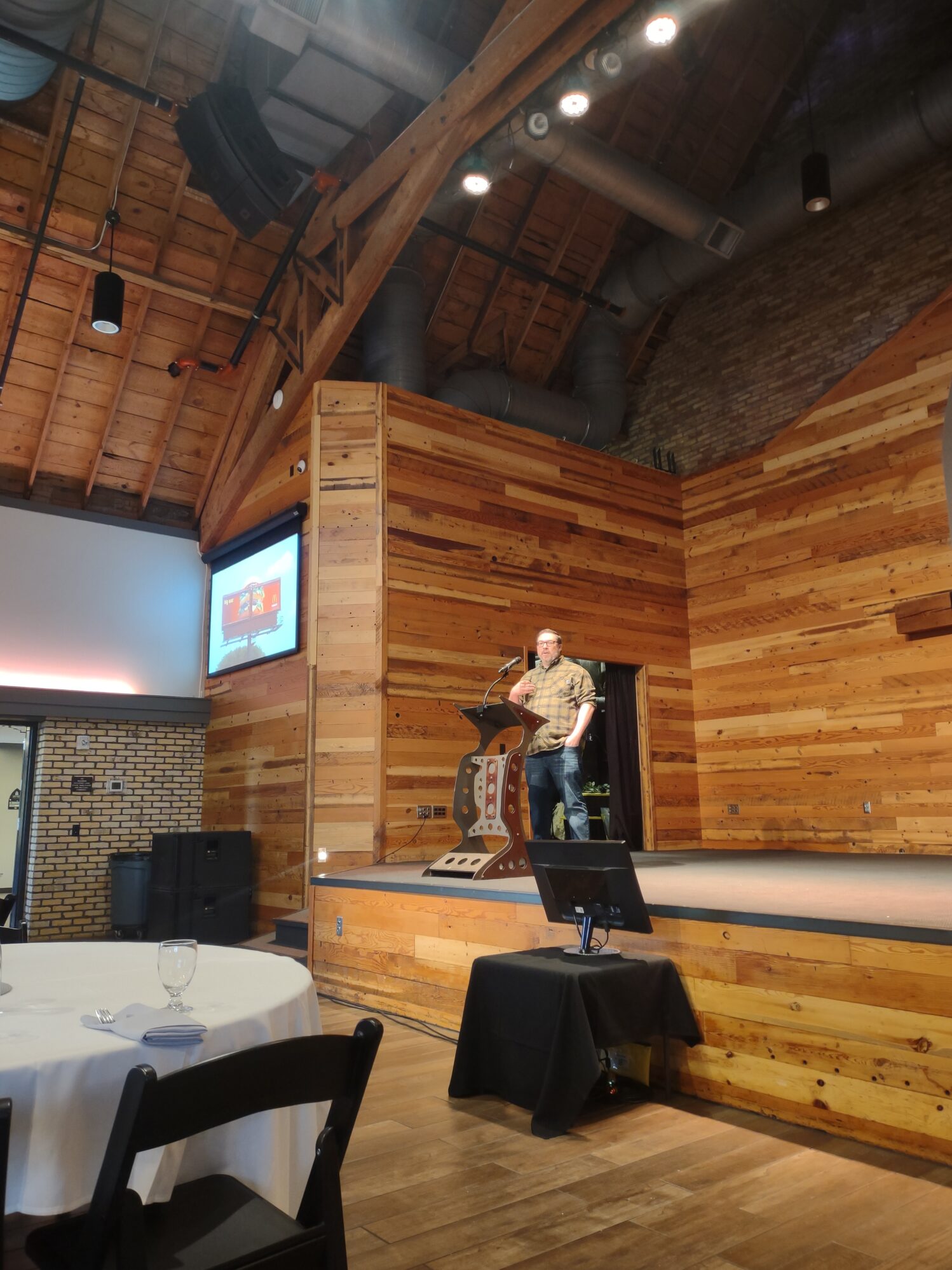 The image shows a man standing on a wooden stage, speaking at a podium in a rustic venue with a high, exposed wooden ceiling and visible ductwork. The walls are made of wooden planks, giving the space a warm, natural feel. There is a projector screen displaying an image on the left side of the stage, and a smaller monitor on a table in front of the stage. In the foreground, there are tables set with white tablecloths, black chairs, and glasses, suggesting a conference or event setup. The atmosphere is professional yet relaxed.