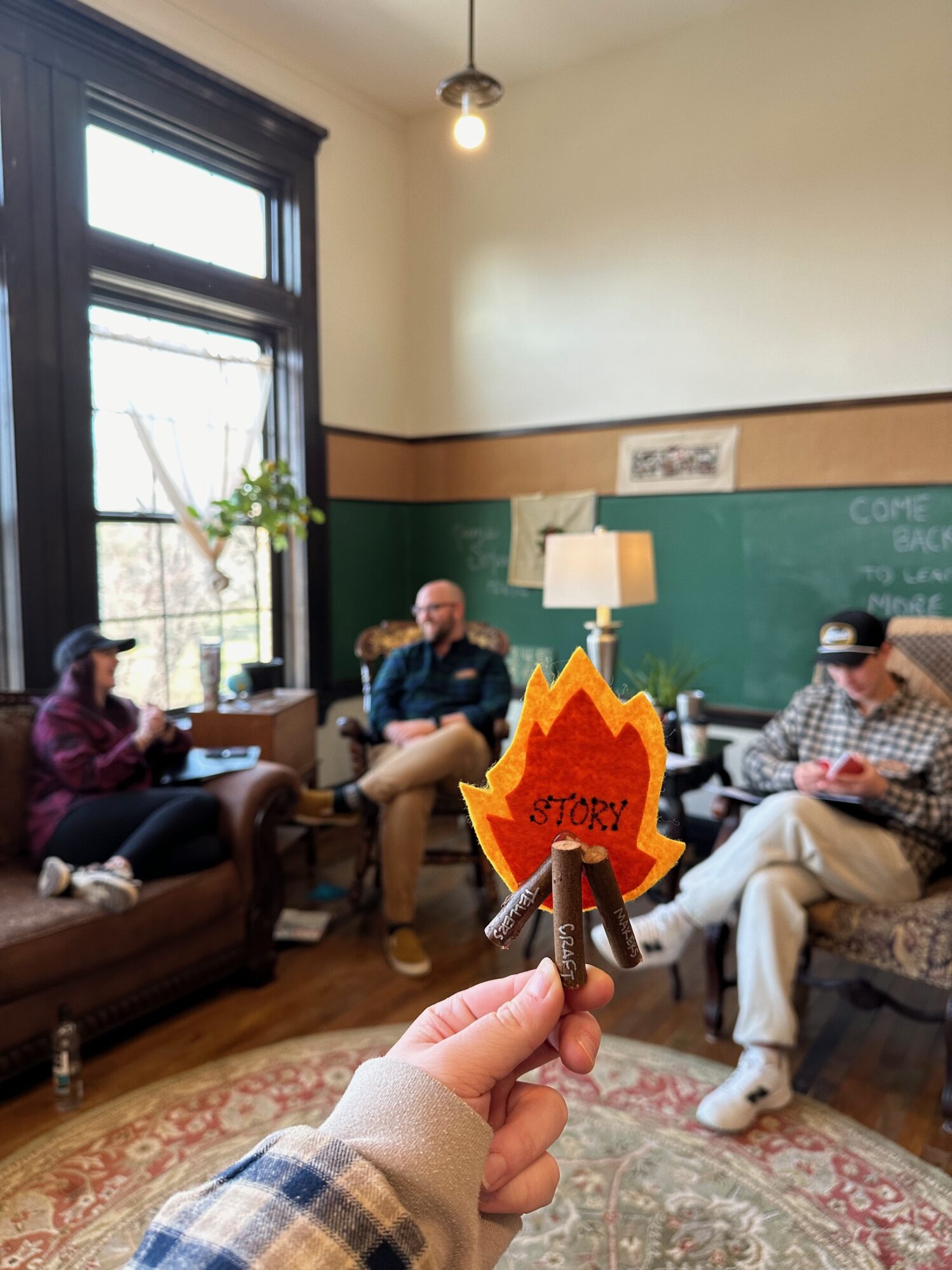 A woman holding an arts and crafts campfire that says “story”. There are three people in the background all wearing flannels. The atmosphere is warm and cheerful.