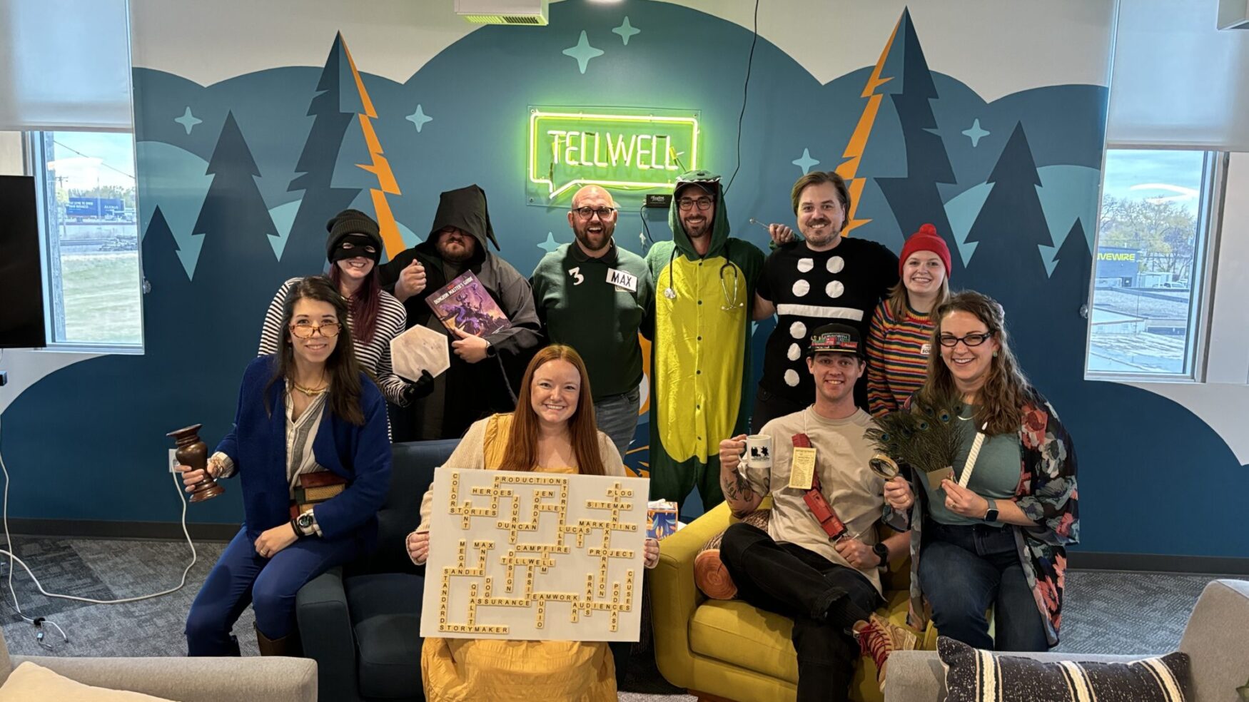 Team photo of Tellwell Story Co. + Studio employees in costume for a themed event. The group is dressed in various creative outfits, including a character in a dinosaur onesie, a villain with a hood, and others holding props like a magnifying glass, a Scrabble board, and a coffee mug. A neon 'Tellwell' sign glows on the wall behind them, with a backdrop of stylized mountain and tree graphics in the office.