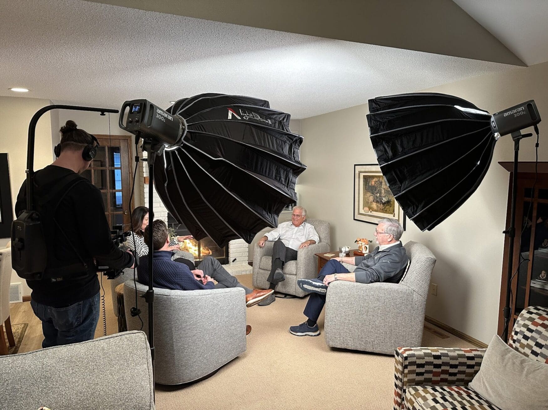 Three men and a woman sit in comfortable chairs around an indoor fireplace. They are lit by two large lights. A man films them as they converse.