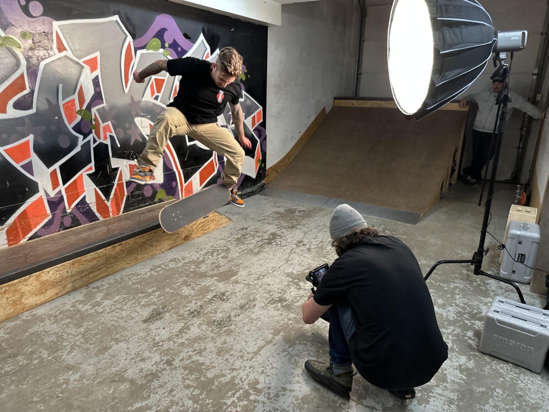 A young man performs a skateboard trick in midair. The moment is captured by a cameraman crouched down in front of him. Colorful graffiti is featured in the background and the scene is lit by a large light.
