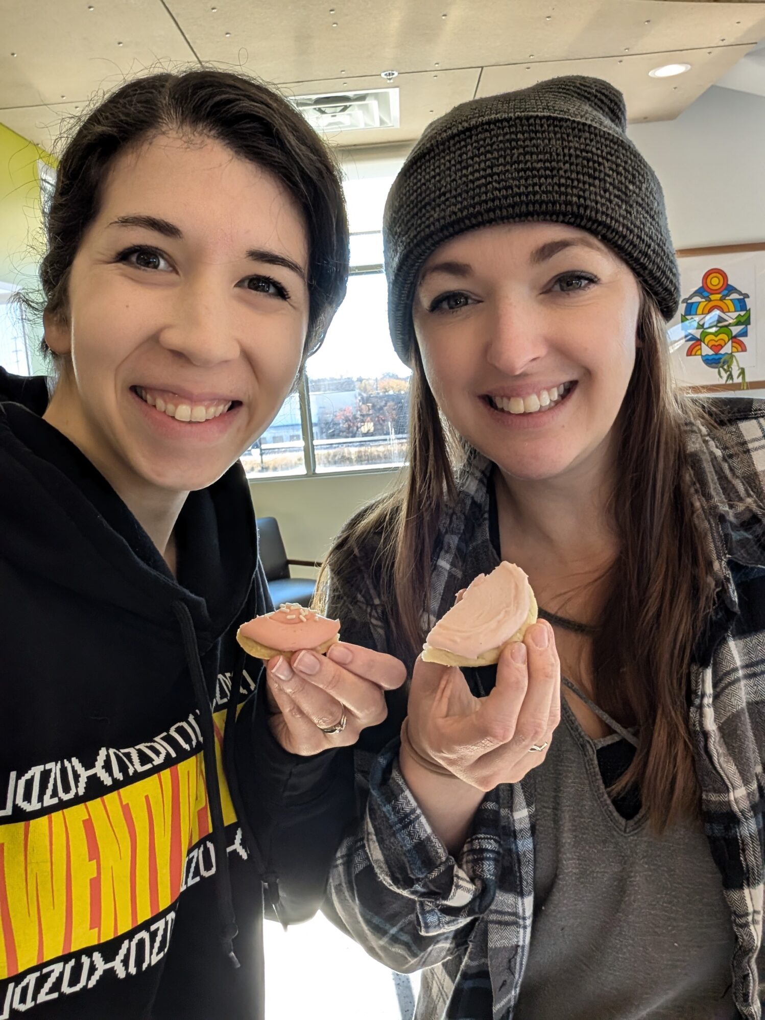 Two smiling women taking a close-up selfie indoors, each holding a frosted sugar cookie with light pink icing. The woman on the left is wearing a black hoodie with yellow and red graphics, while the woman on the right is wearing a gray beanie and a plaid shirt layered over a gray top. Behind them, there is a window with natural light and colorful artwork on the wall.