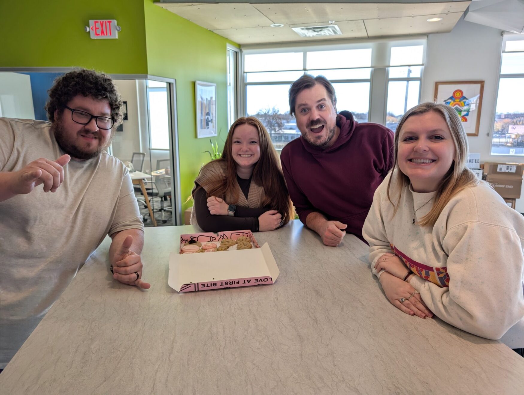 Four smiling people gathered around a table in a brightly lit indoor space. In the center of the table is an open pink box of assorted cookies. The person on the left, wearing a beige shirt, is giving a thumbs-up, while the others lean in cheerfully. The background features green and white walls, a window, and a colorful artwork on the wall.