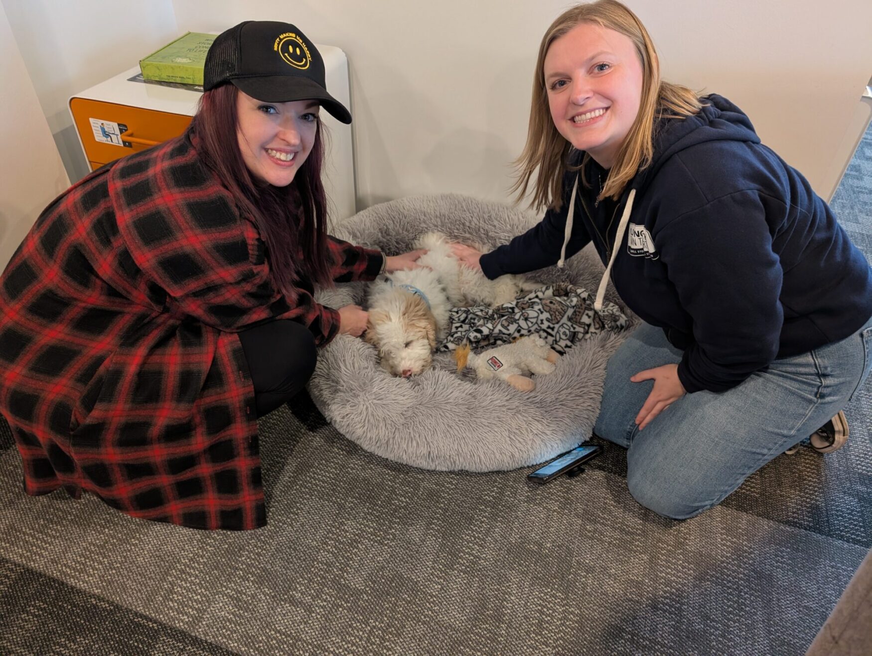 Two women pet a medium sized dog laying in a dog bed. One woman has a red plaid shirt and black hat; the other is wearing a dark blue hoodie with blue jeans.