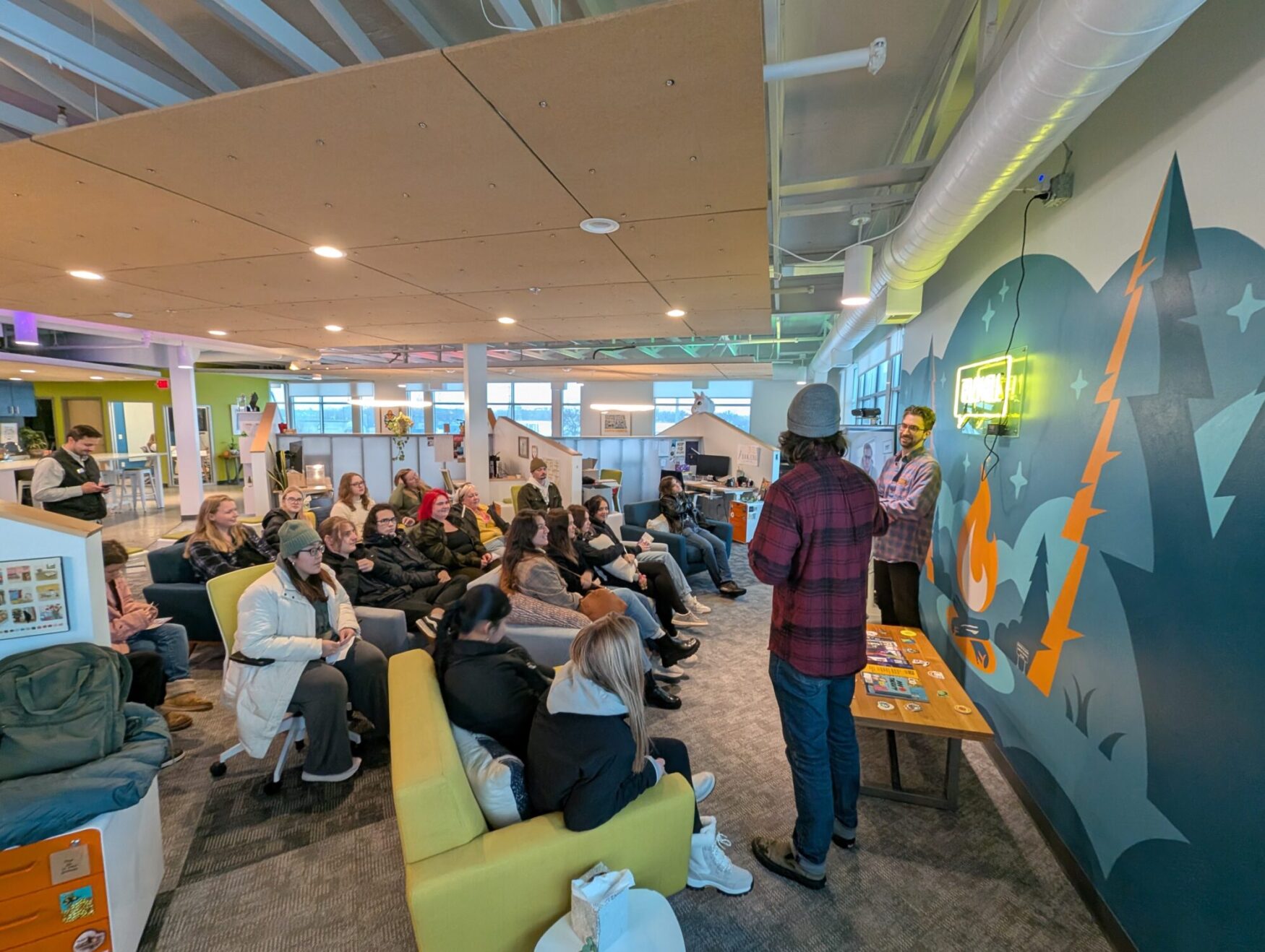 A group of about 10-20 college age students sits together watching a presentation in a colorful office space. Two in person male presenters and one on a TV screen in the late 20’s to early 30’s stand in front of the students.