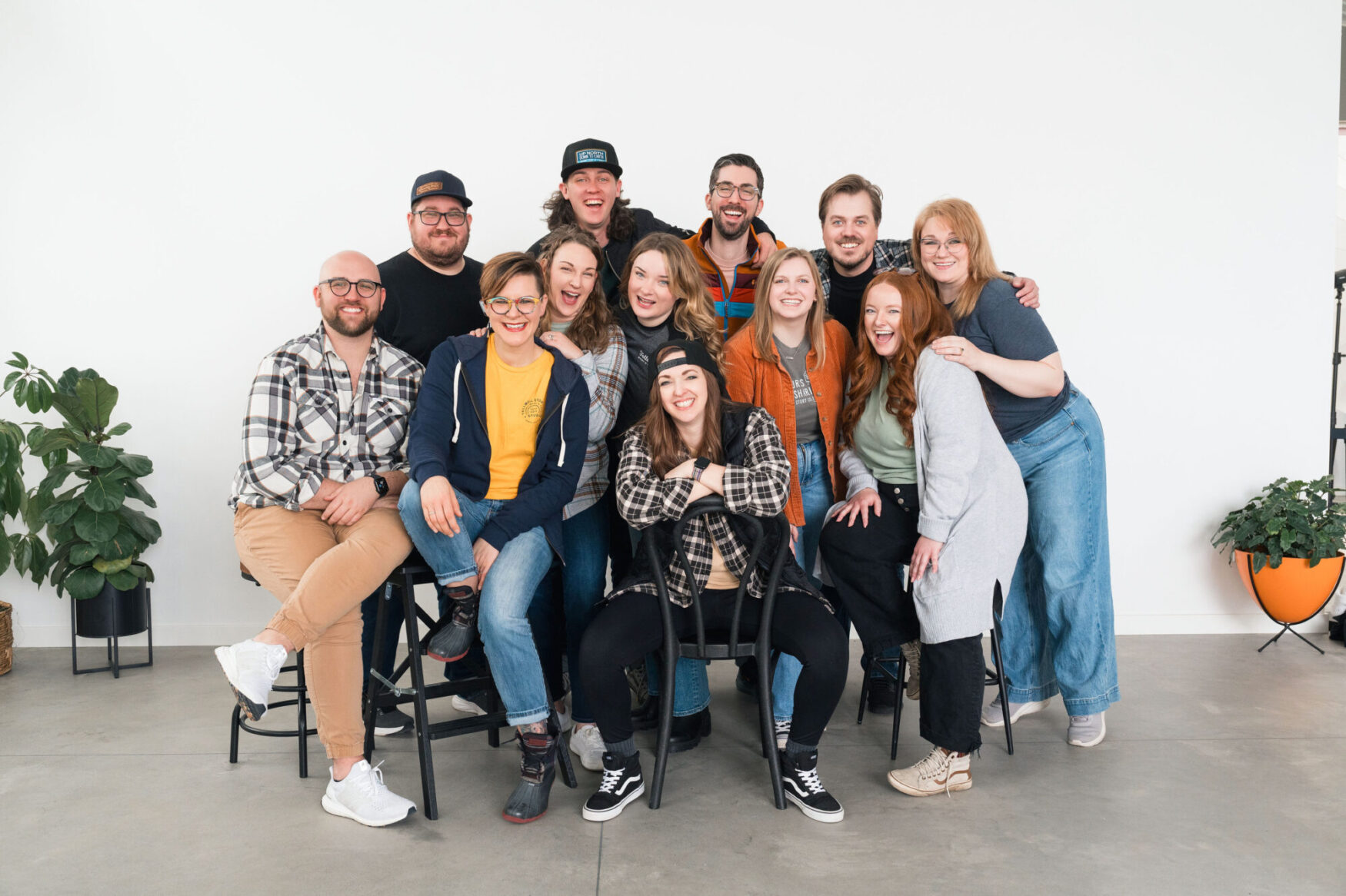 Group photo of 13 smiling team members from Tellwell Story Co. + Studio in a well-lit studio space. Everyone is casually dressed, sitting and standing close together, with a relaxed and friendly vibe. Two potted plants on either side add a touch of greenery to the setting.