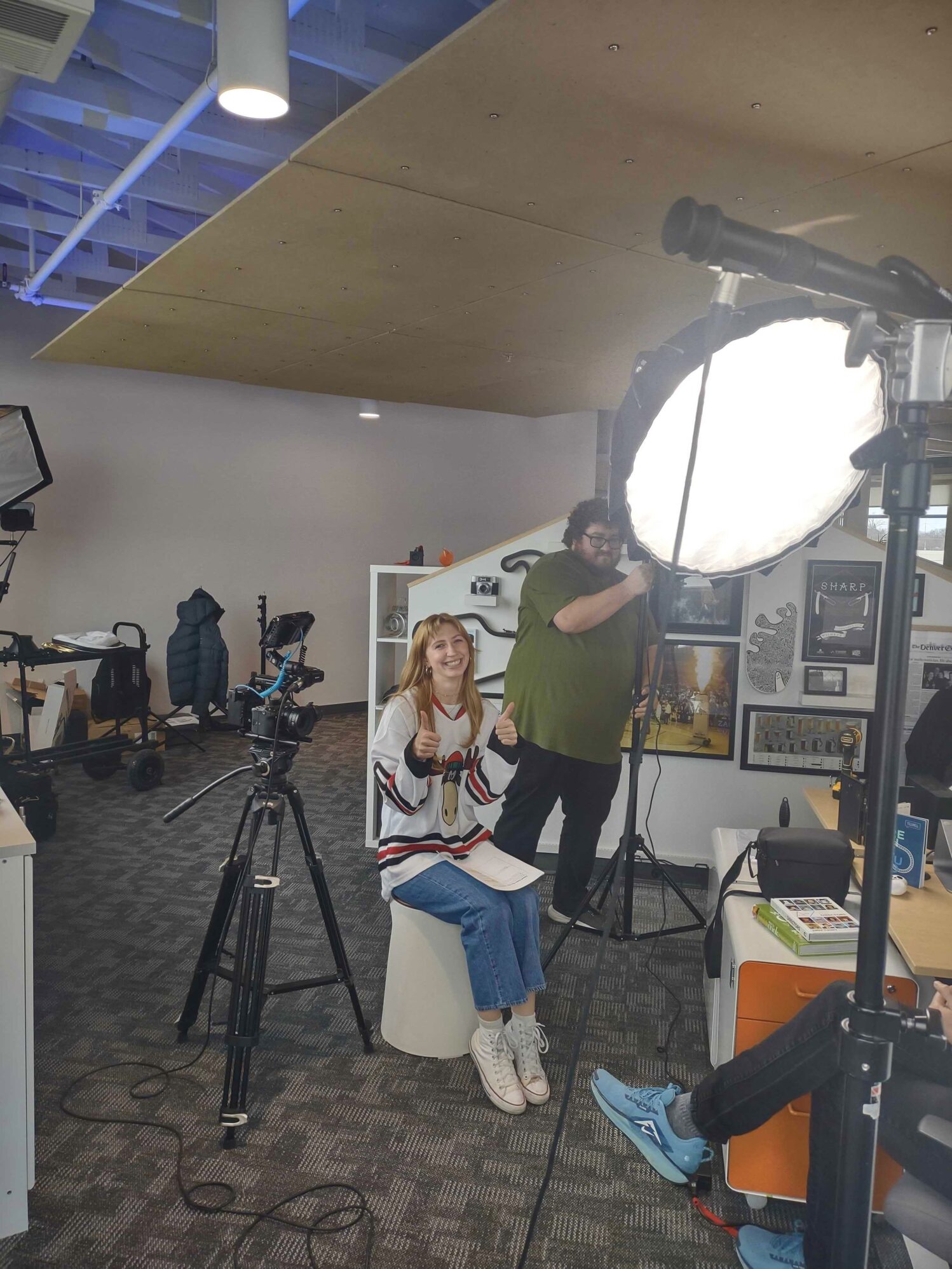 Behind-the-scenes photo of a casual video shoot in an office setting. A smiling woman in a hockey jersey gives two thumbs up while seated in front of a camera on a tripod, surrounded by lighting equipment. A crew member adjusts the lighting in the background.