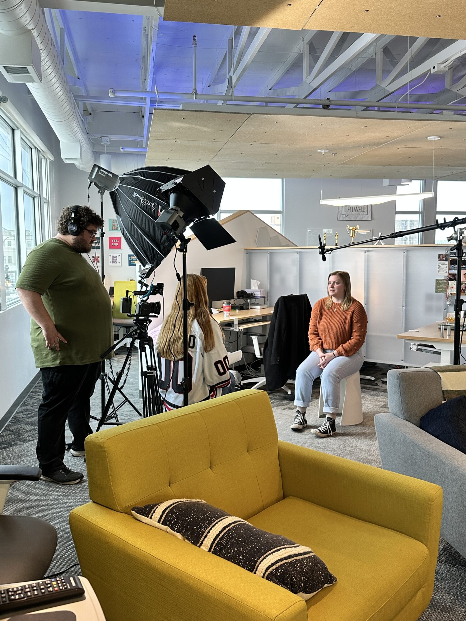 Behind-the-scenes photo of a video interview in a bright office space. A woman sits on a stool in front of a camera and lighting setup, while a crew member adjusts the equipment. Another person wearing a hockey jersey is partially visible, seated with their back to the camera. A yellow couch with a striped pillow is in the foreground.