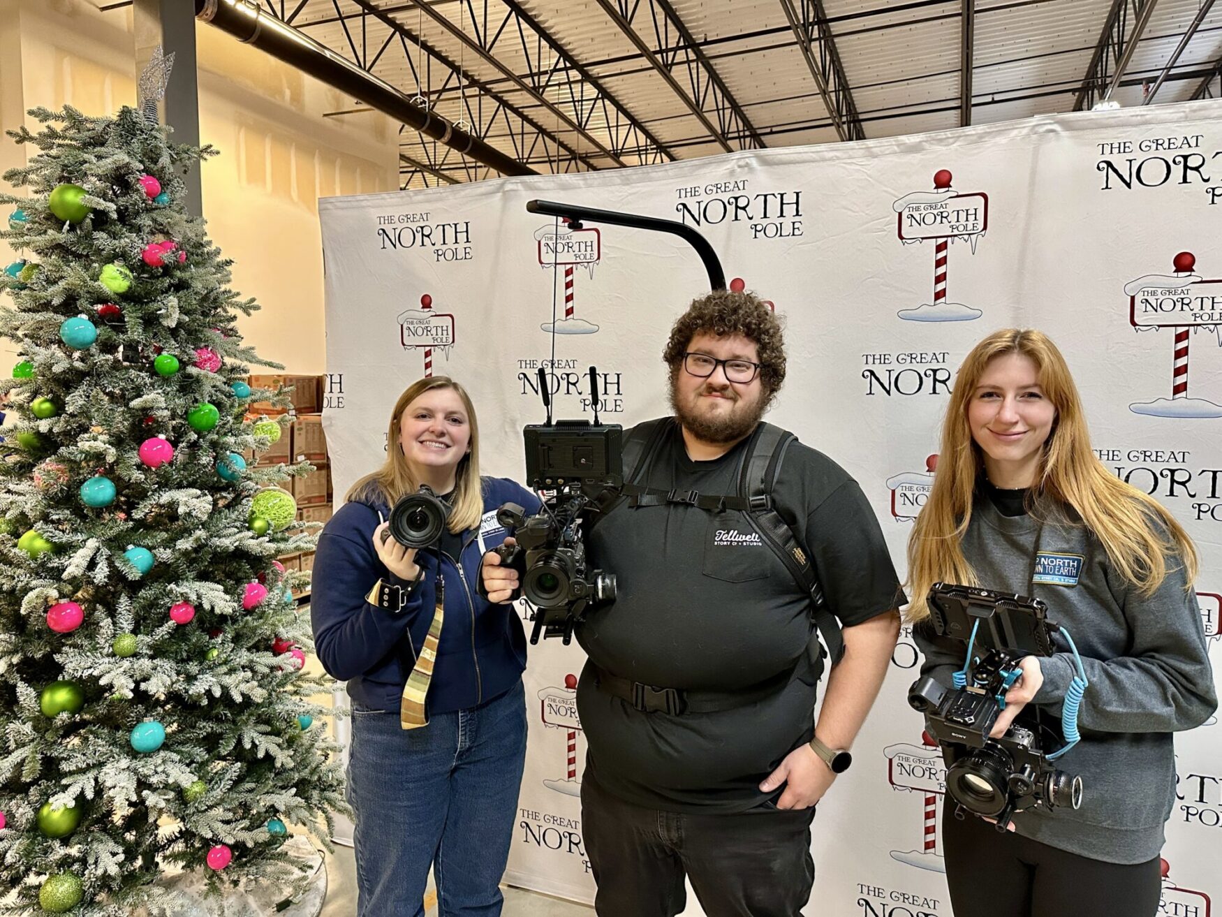 The image shows three people holding professional camera equipment, standing in front of a festive backdrop that says "The Great North Pole." On the left is a Christmas tree decorated with colorful ornaments, including green, pink, and blue ones. The setting appears to be an indoor event space with a holiday theme.